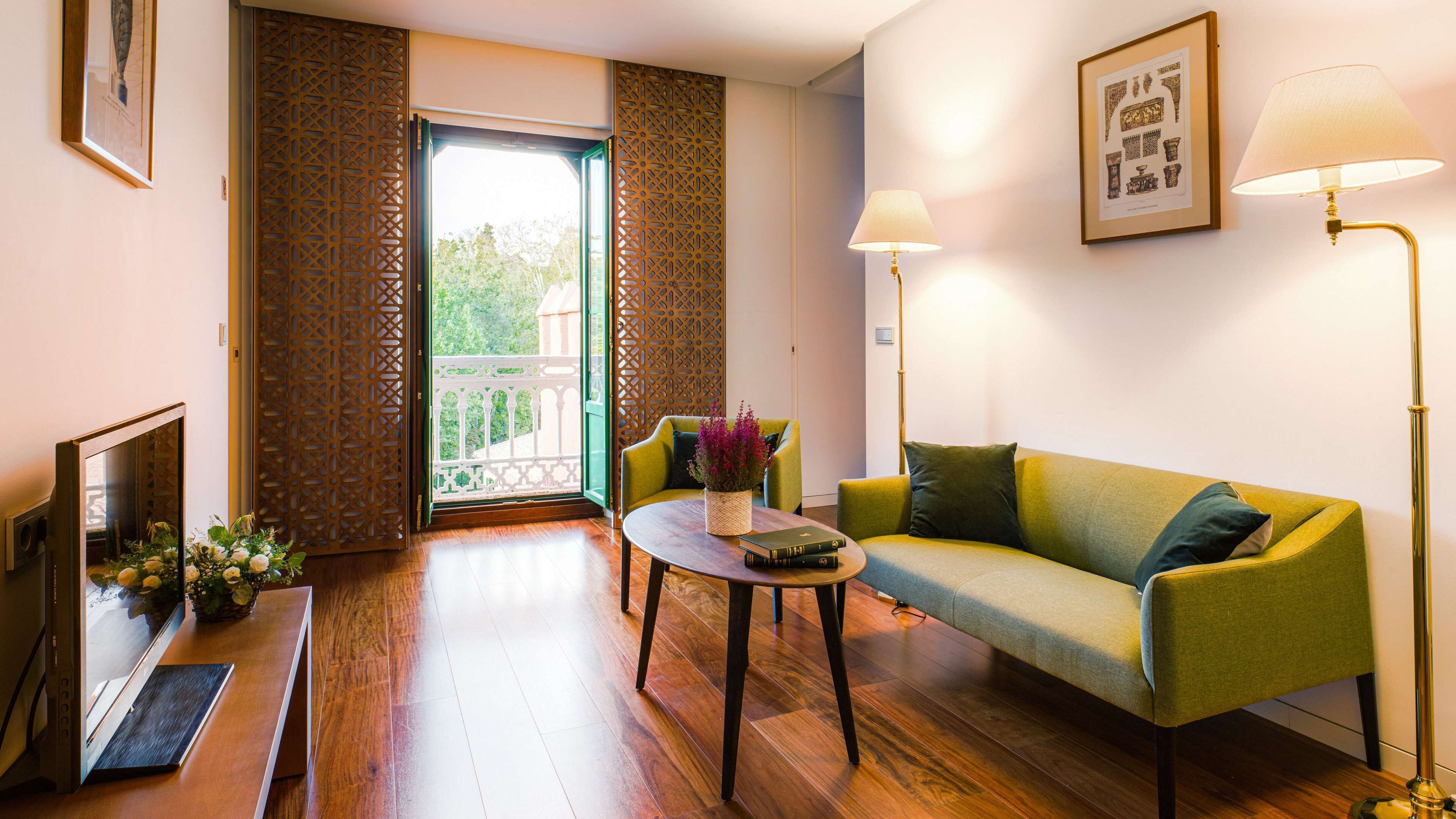 A living room with a green sofa, a coffee table with books and flowers, a TV, and a balcony with wooden lattice doors.