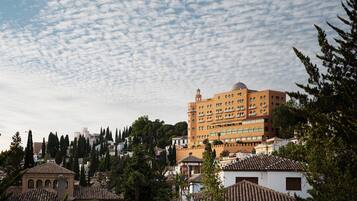 Un edificio grande de varios pisos con una torre destacada, rodeado de edificios y árboles más pequeños.