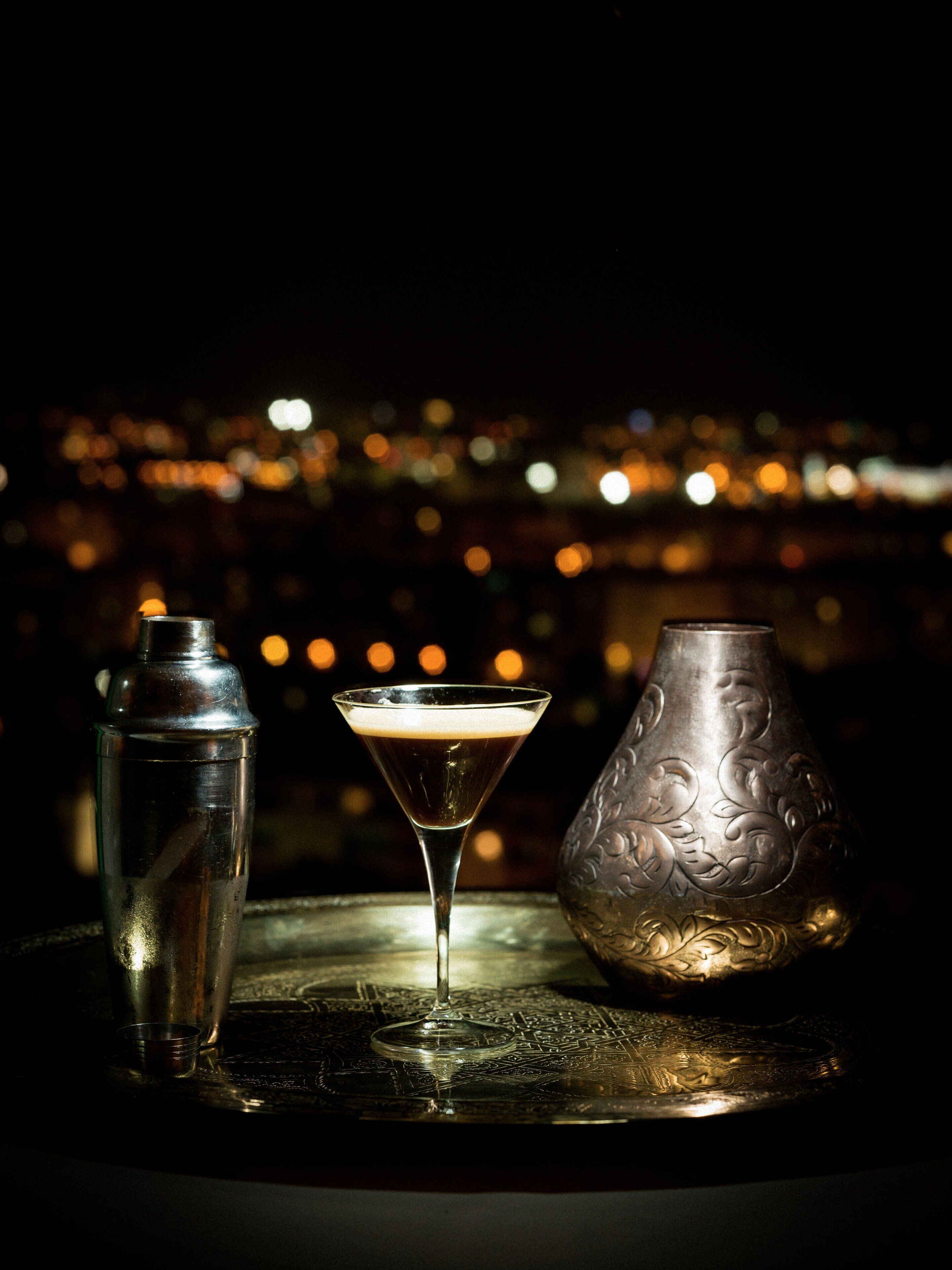 A cocktail glass, a metal shaker, and a decorative vase on a table with a cityscape background.