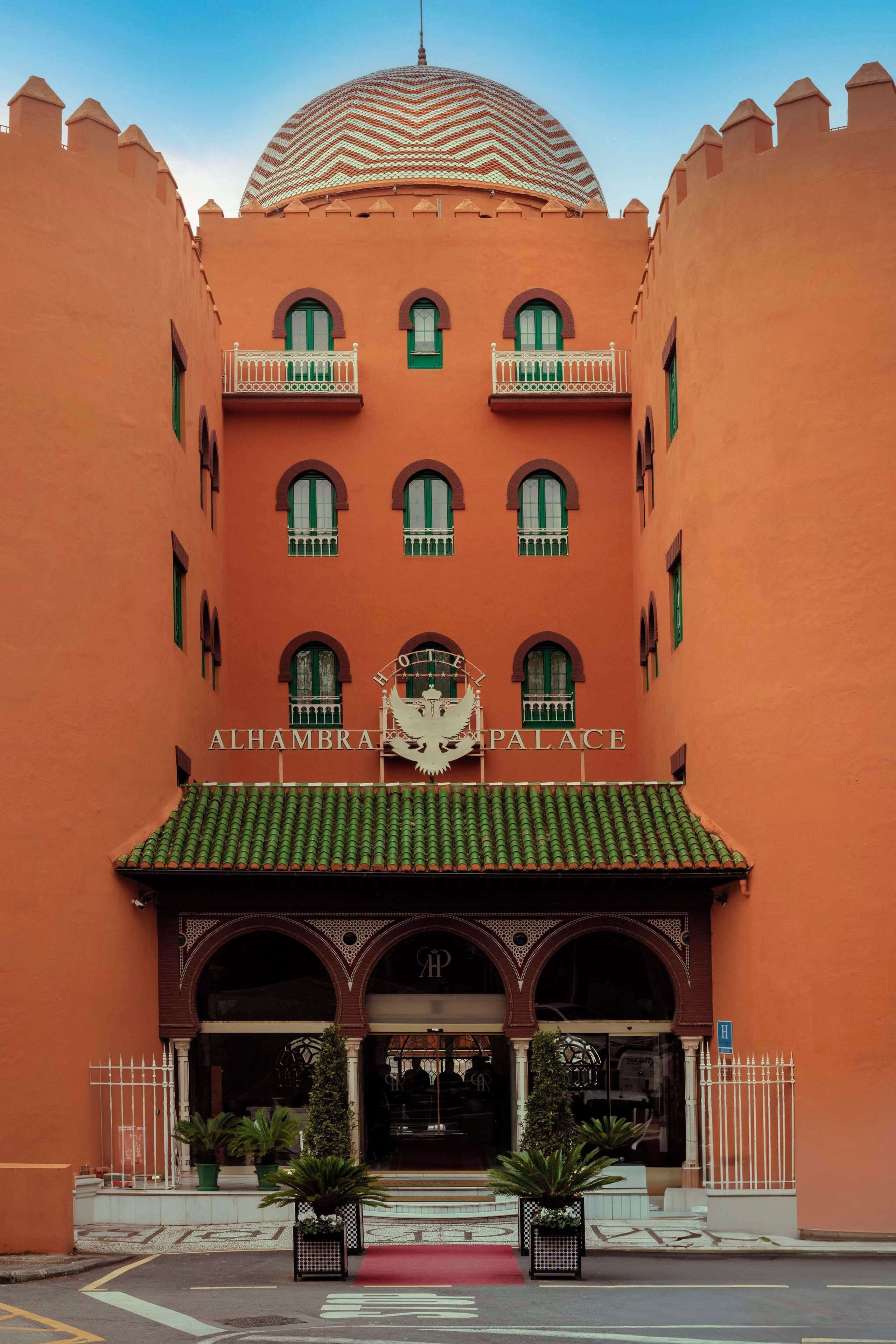 A large, orange building with a central dome and multiple balconies. The entrance features a red carpet and a sign reading "ALHAMBRA PALACE".
