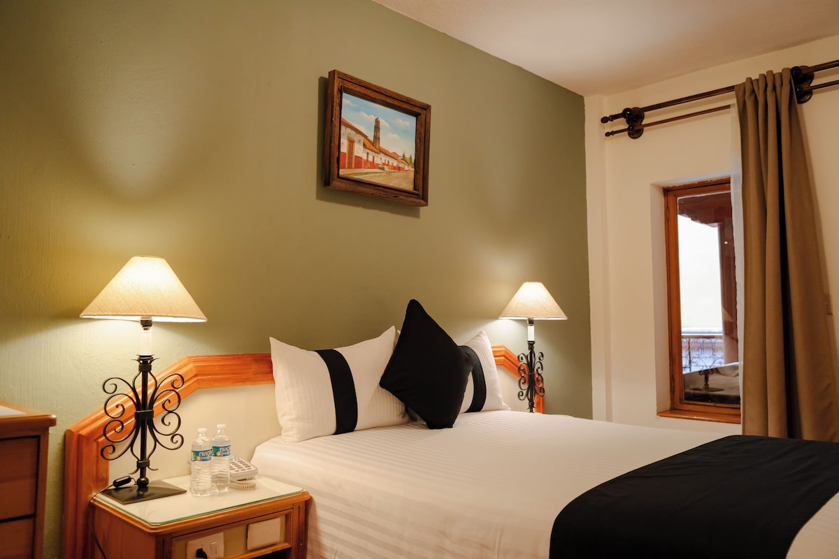 A hotel room with a bed, two bedside lamps, a nightstand, a painting on the wall, and a window with curtains.