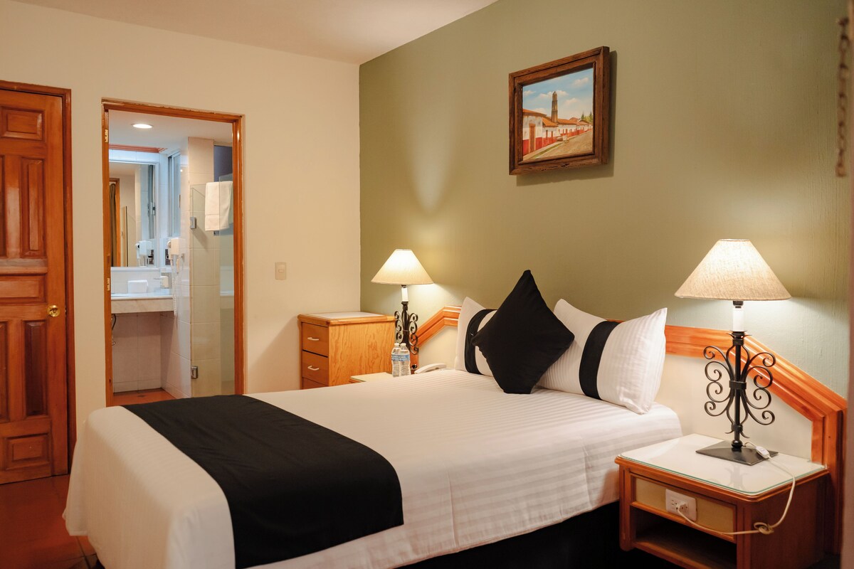 A hotel room with a bed, two bedside lamps, a nightstand, a wooden headboard, a painting on the wall, and a bathroom visible through an open door.
