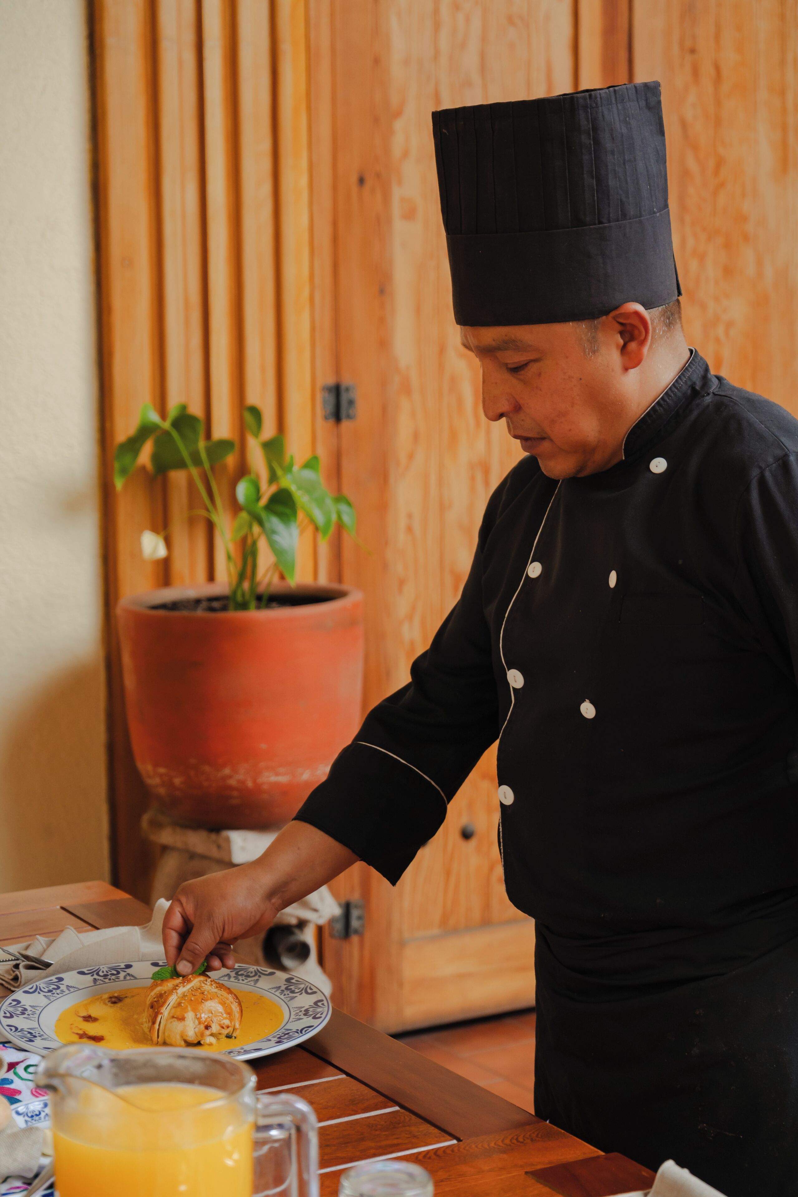 A chef in a black uniform with a tall hat, preparing food on a plate with a yellow sauce.