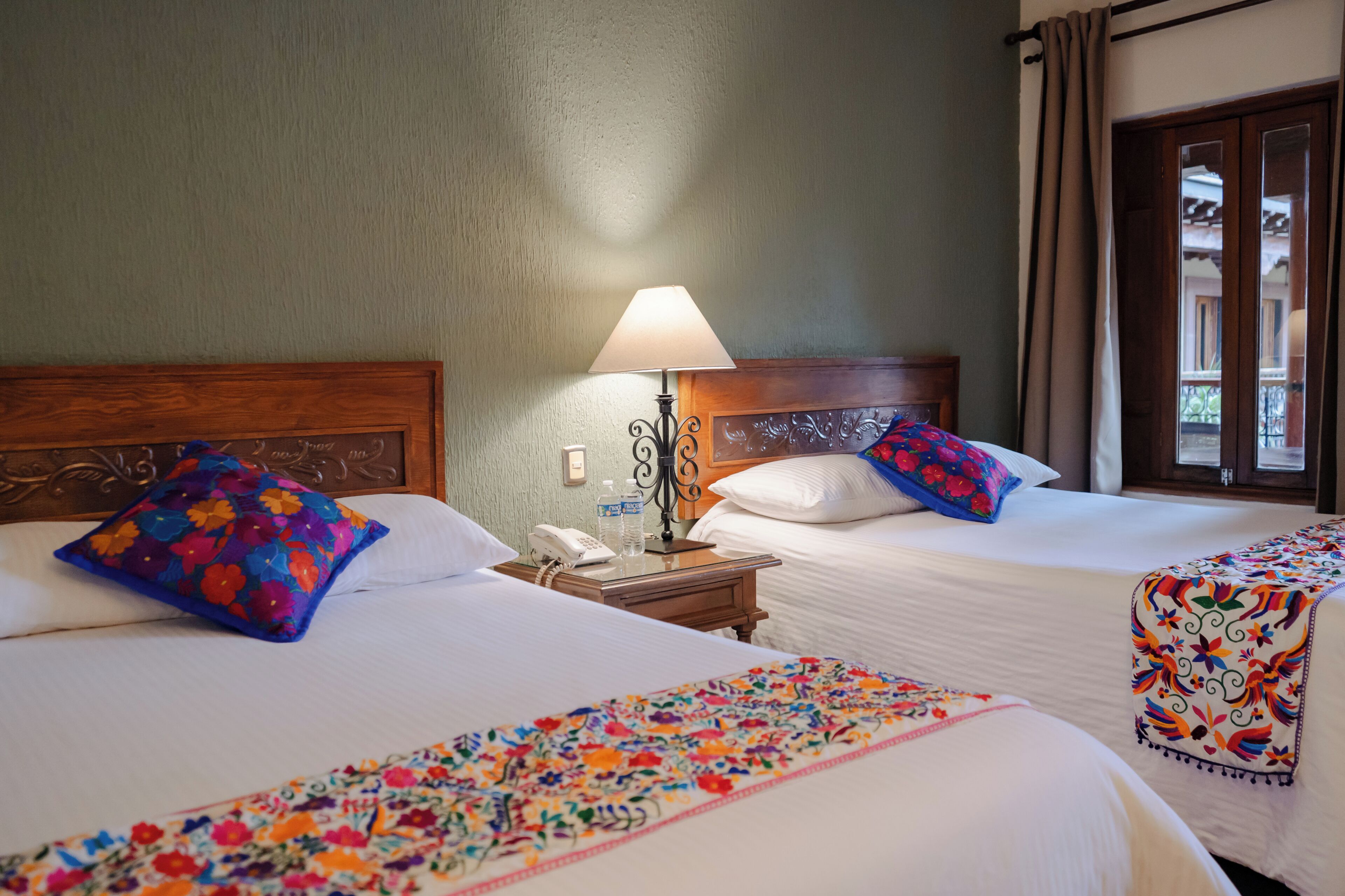 Two beds with floral bedspreads, a bedside table with a lamp, and a window with curtains.