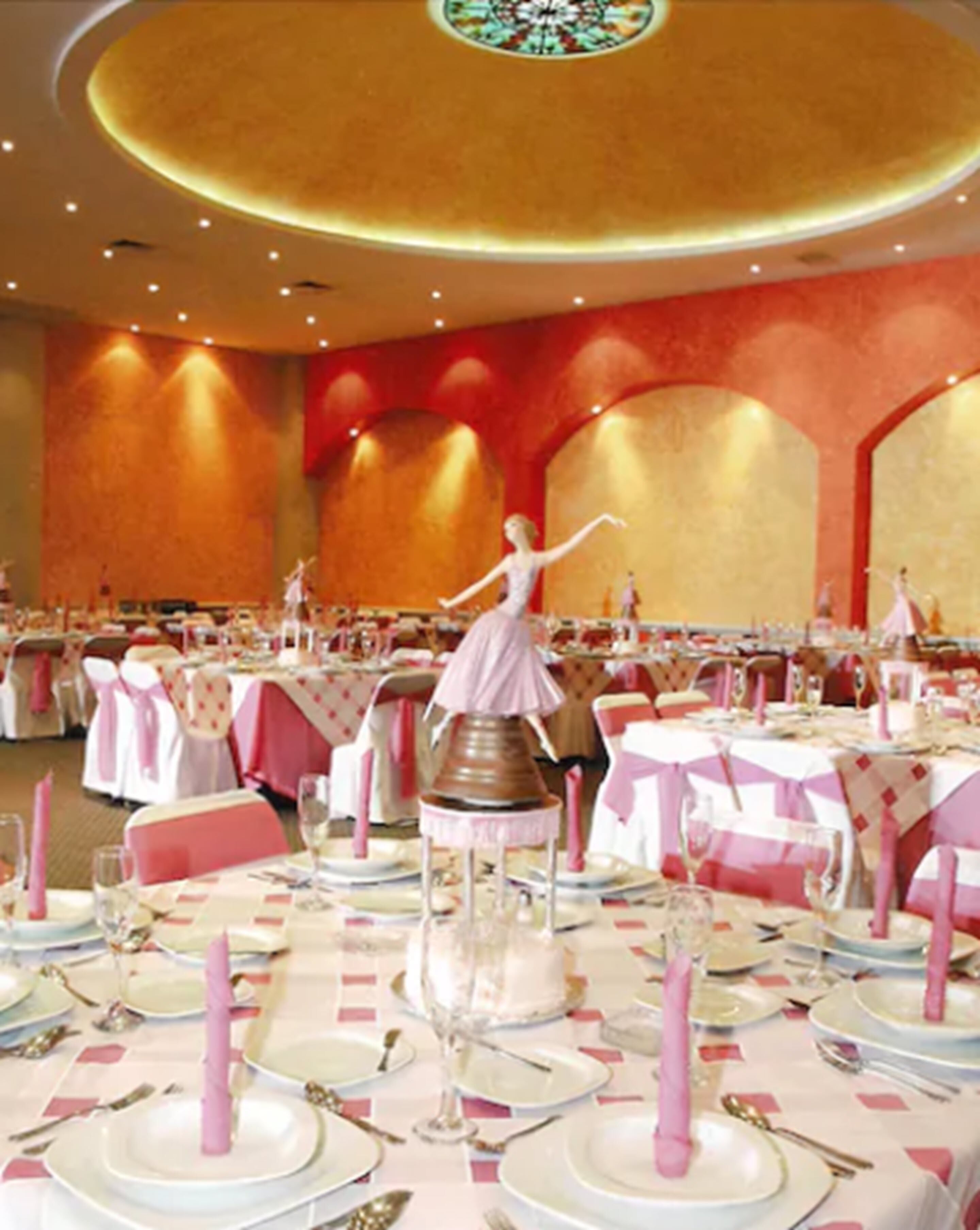Un salón de banquetes con mesas redondas dispuestas para un evento formal, con una decoración central y detalles en rosa.