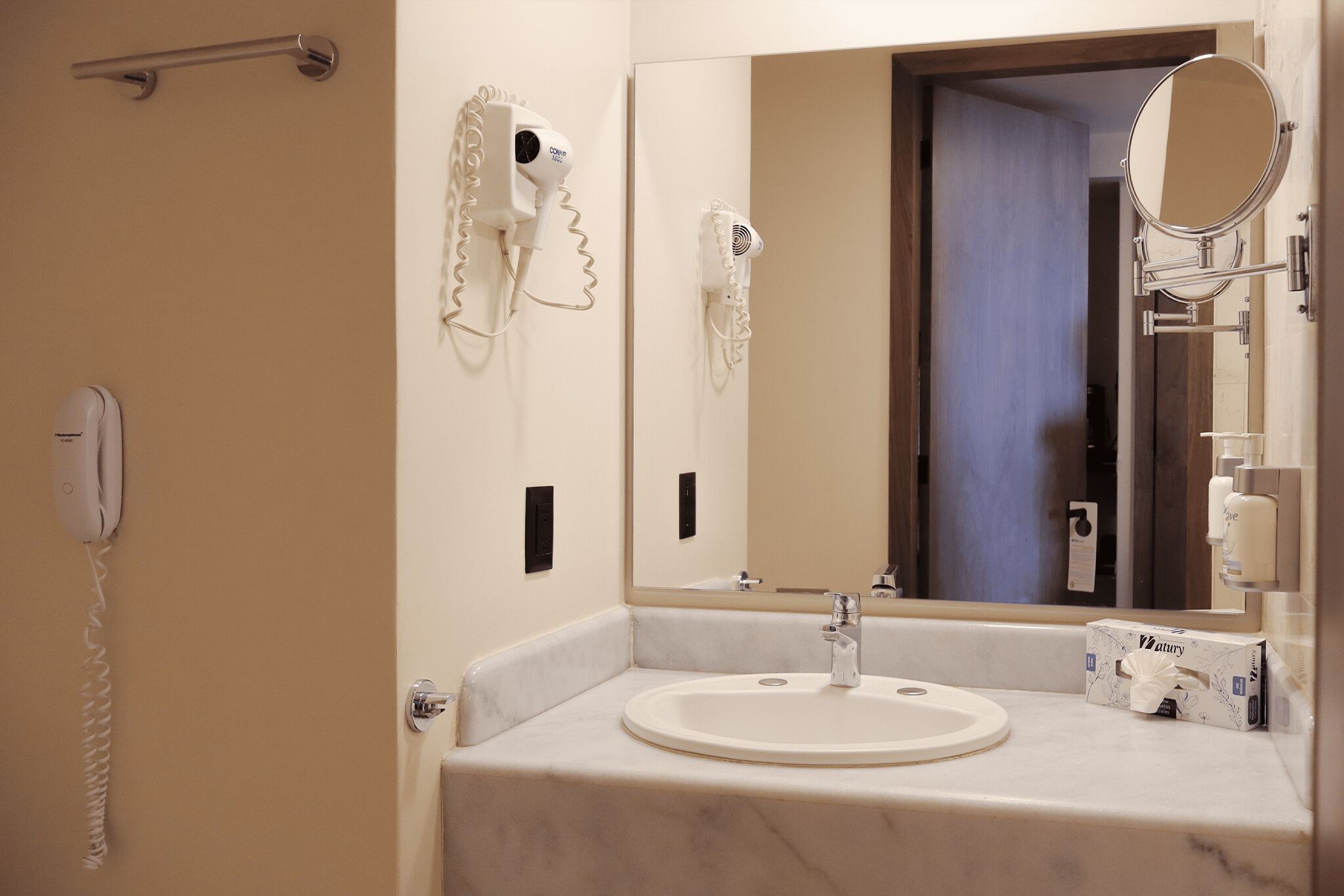 A bathroom with a sink, a hairdryer, a mirror, a telephone, and a box of tissues.