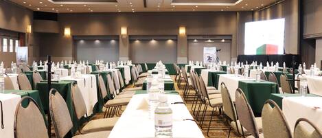 A conference room with rows of tables, chairs, and water bottles, equipped with a projector screen.