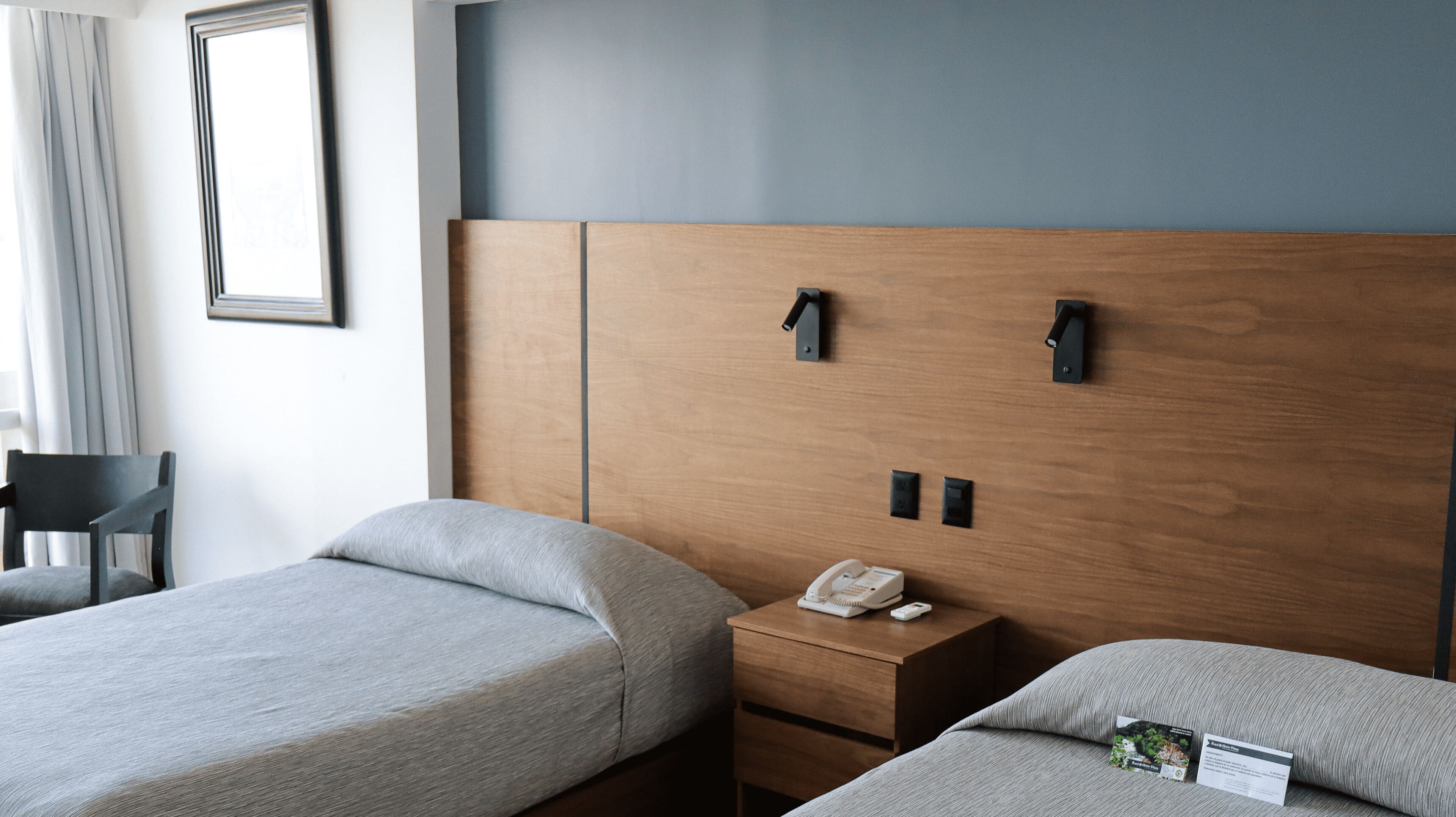 Habitación de hotel con dos camas individuales, cabecera de madera, mesita de noche con teléfono y una silla.