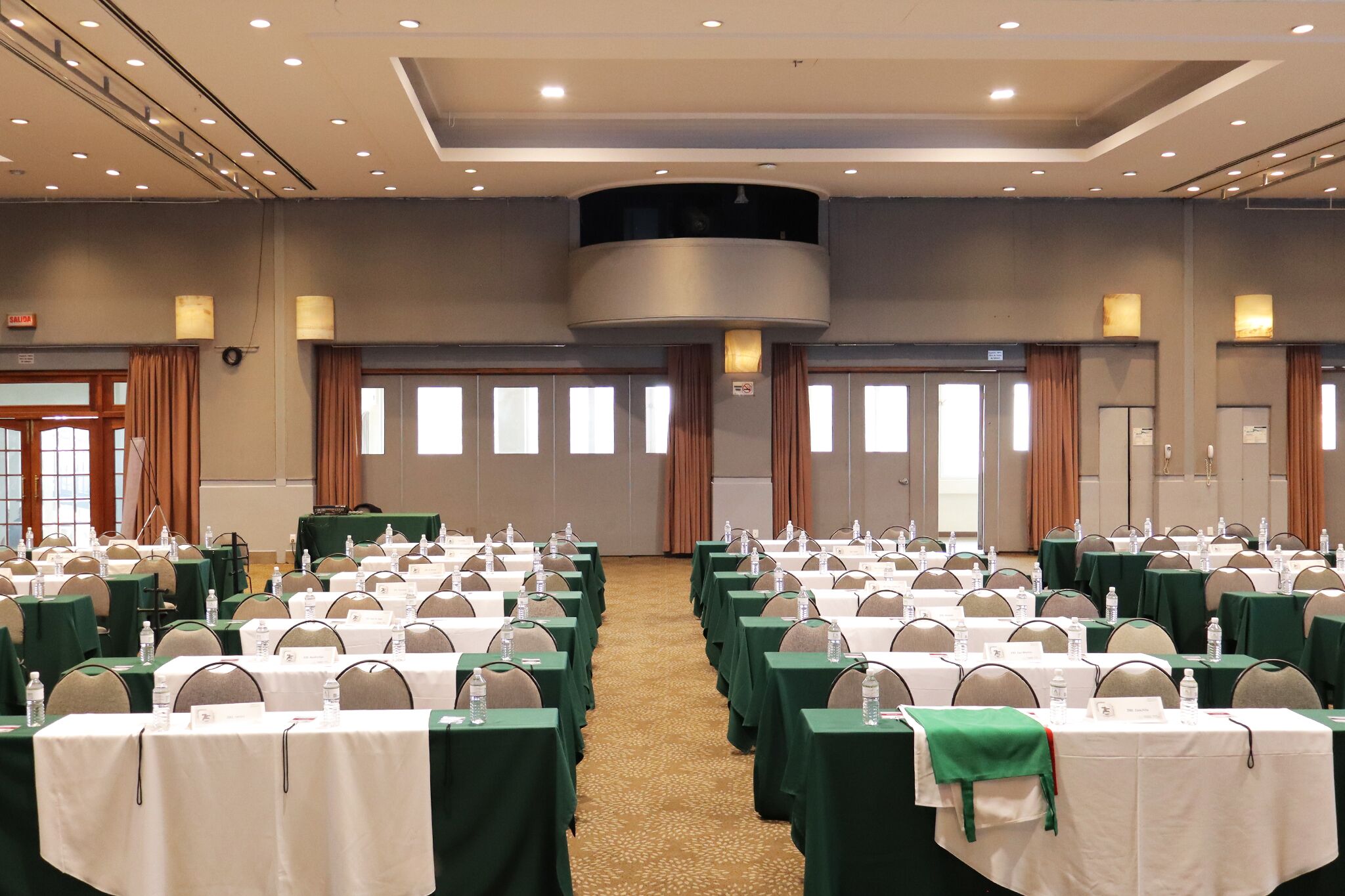 Un salón de conferencias con filas de mesas, sillas y botellas de agua, con piso alfombrado y techo alto con iluminación empotrada.
