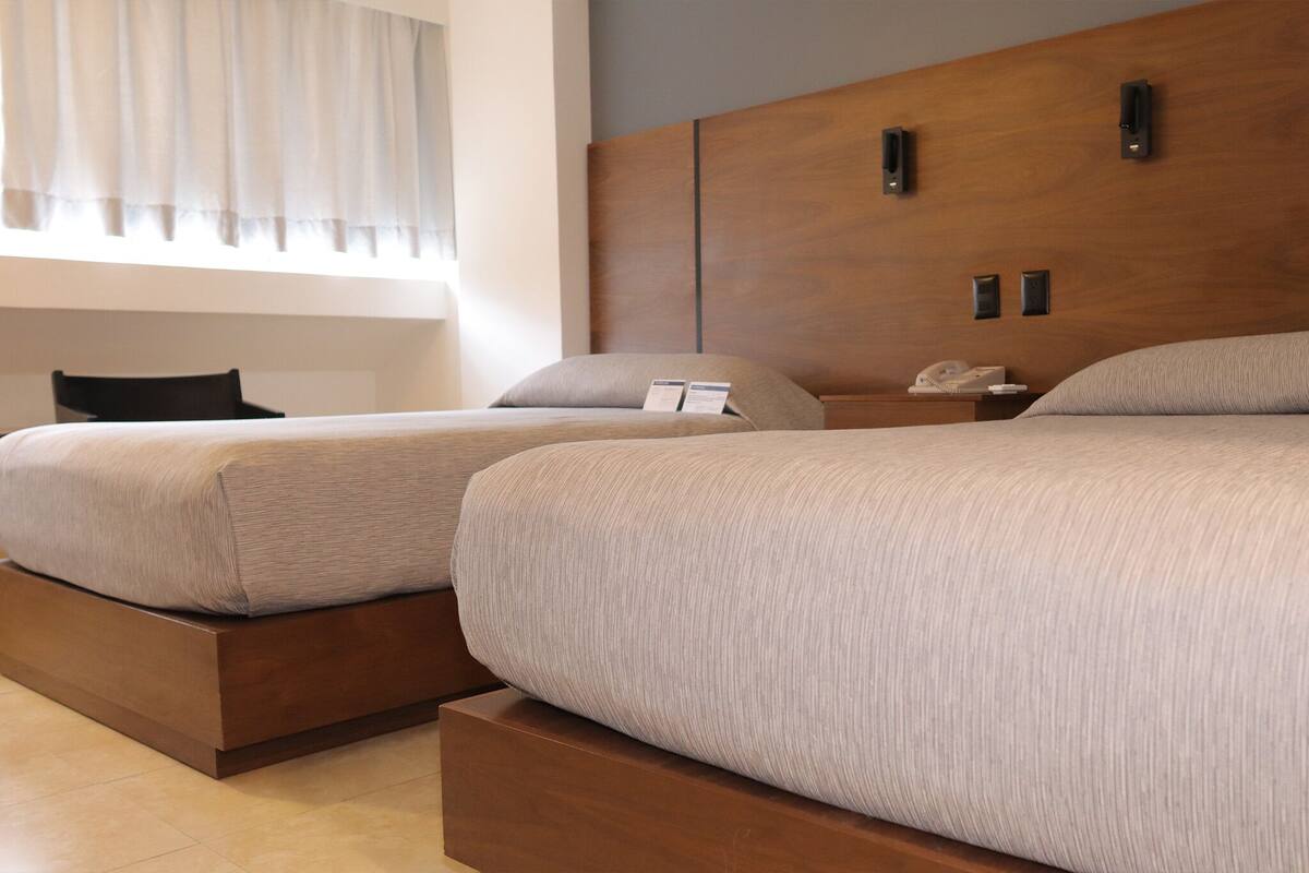 Une chambre d’hôtel avec deux lits, une tête de lit en bois, des tables de chevet avec des lampes, une chaise et une fenêtre avec des rideaux.