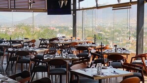 Un restaurant amb taules preparades per sopar, un gran finestral que ofereix vistes a la ciutat i un televisió de pantalla plana penjada a la paret.