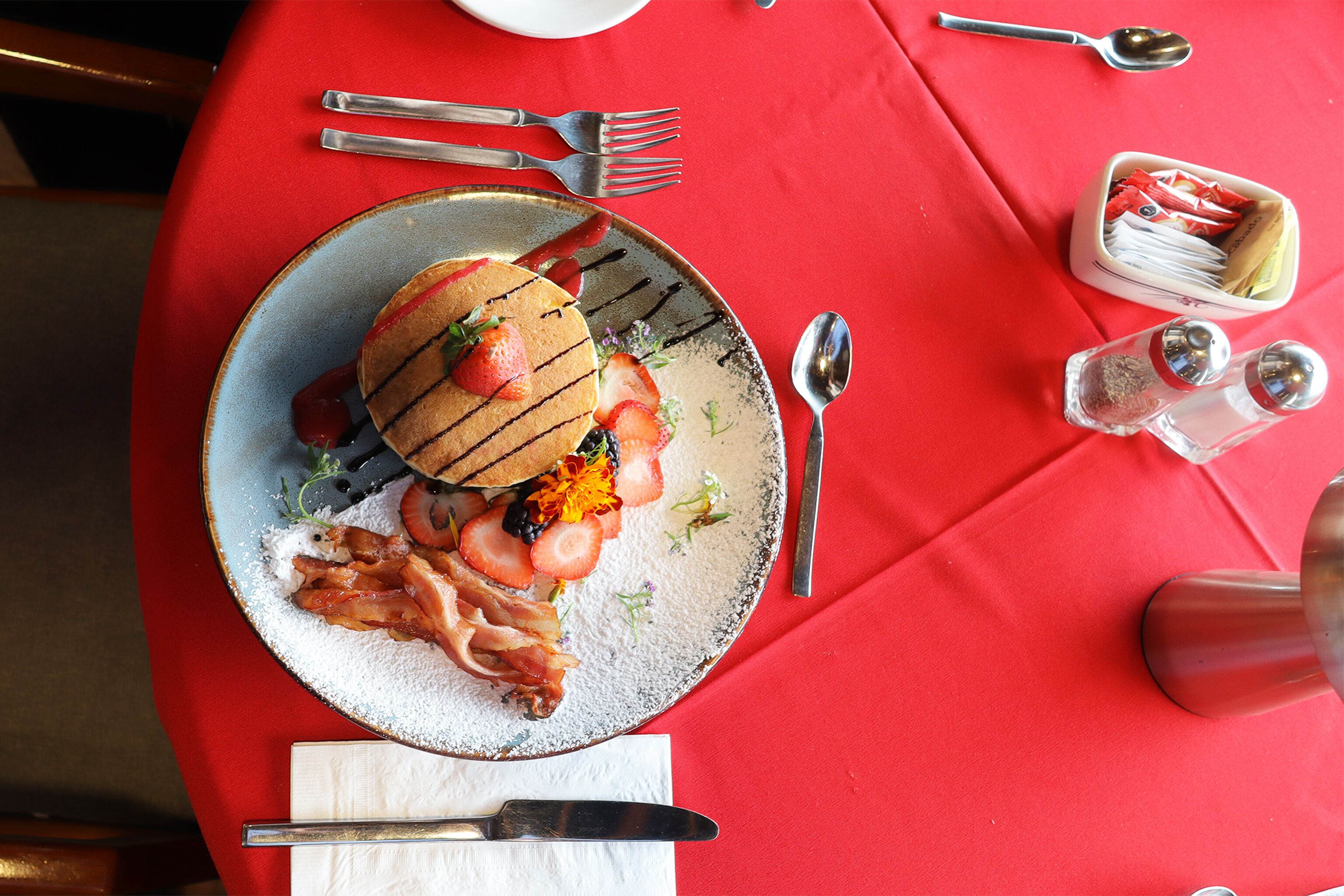 Ein Frühstücksteller mit Pfannkuchen, Erdbeeren und Speck auf einer roten Tischdecke.