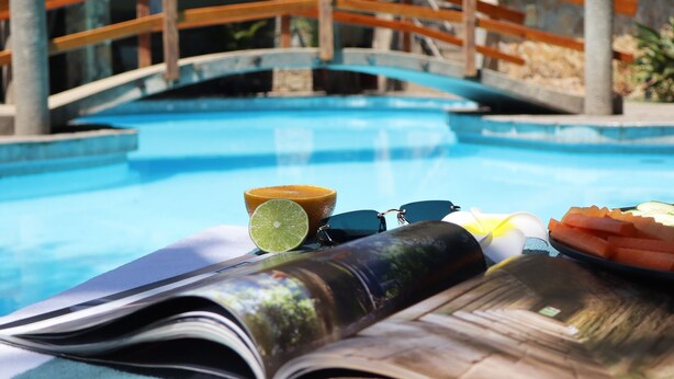 Escena a la piscina amb un pont de fusta, un llibre, meitat d'un llimó, ulleres de sol i un plat de verdures tallades.