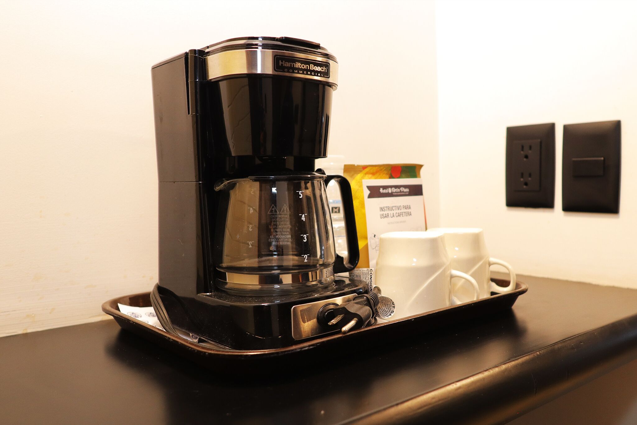 A black Hamilton Beach coffee maker on a tray with two white cups and a coffee pod.