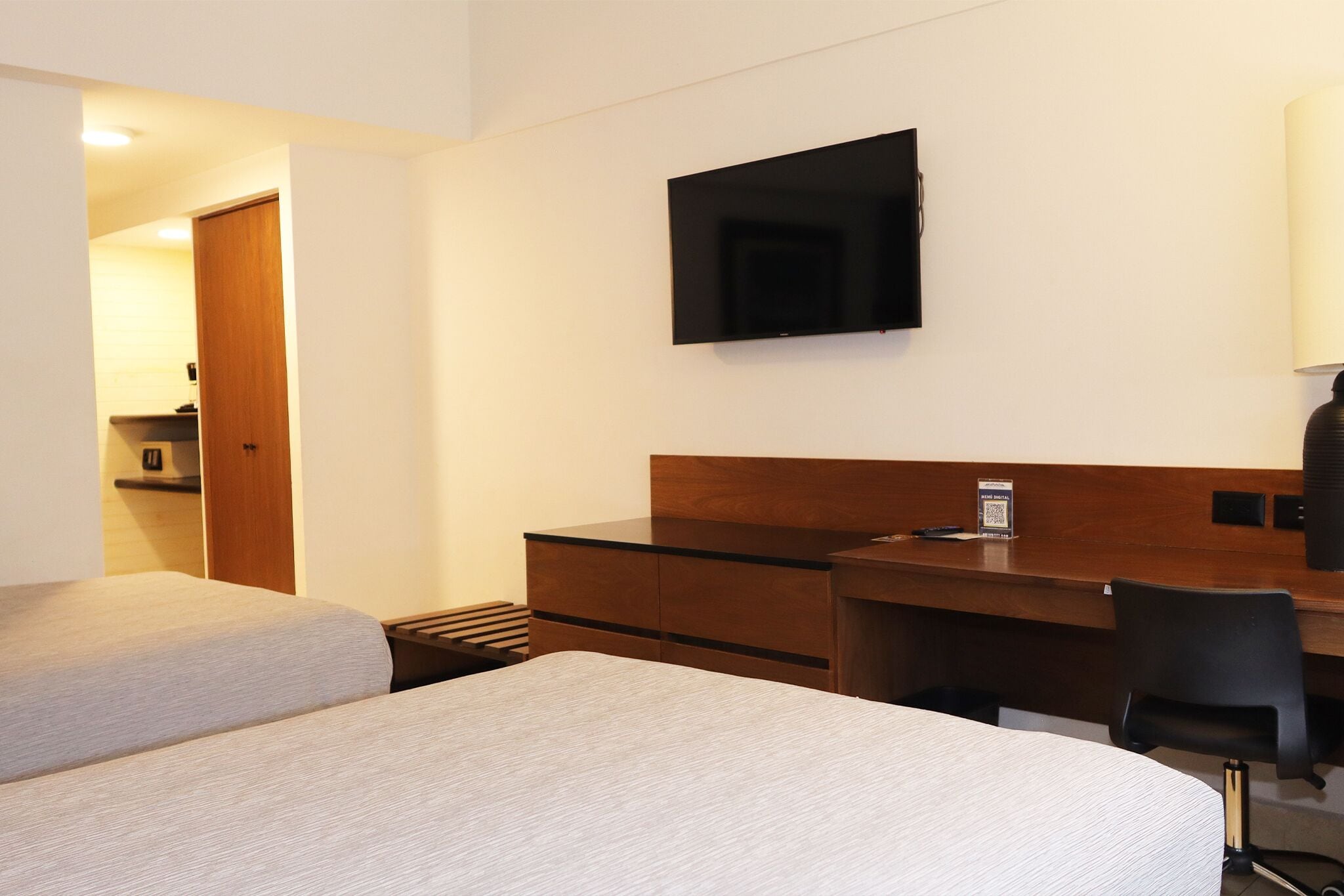 A hotel room with two beds, a desk, a chair, a flat-screen TV, and a small kitchenette.
