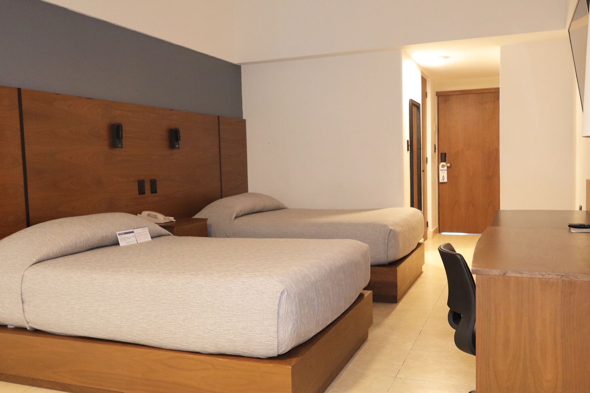 Ein Hotelzimmer mit zwei Betten, einem hölzernen Kopfteil, einem Schreibtisch und einer Tür.