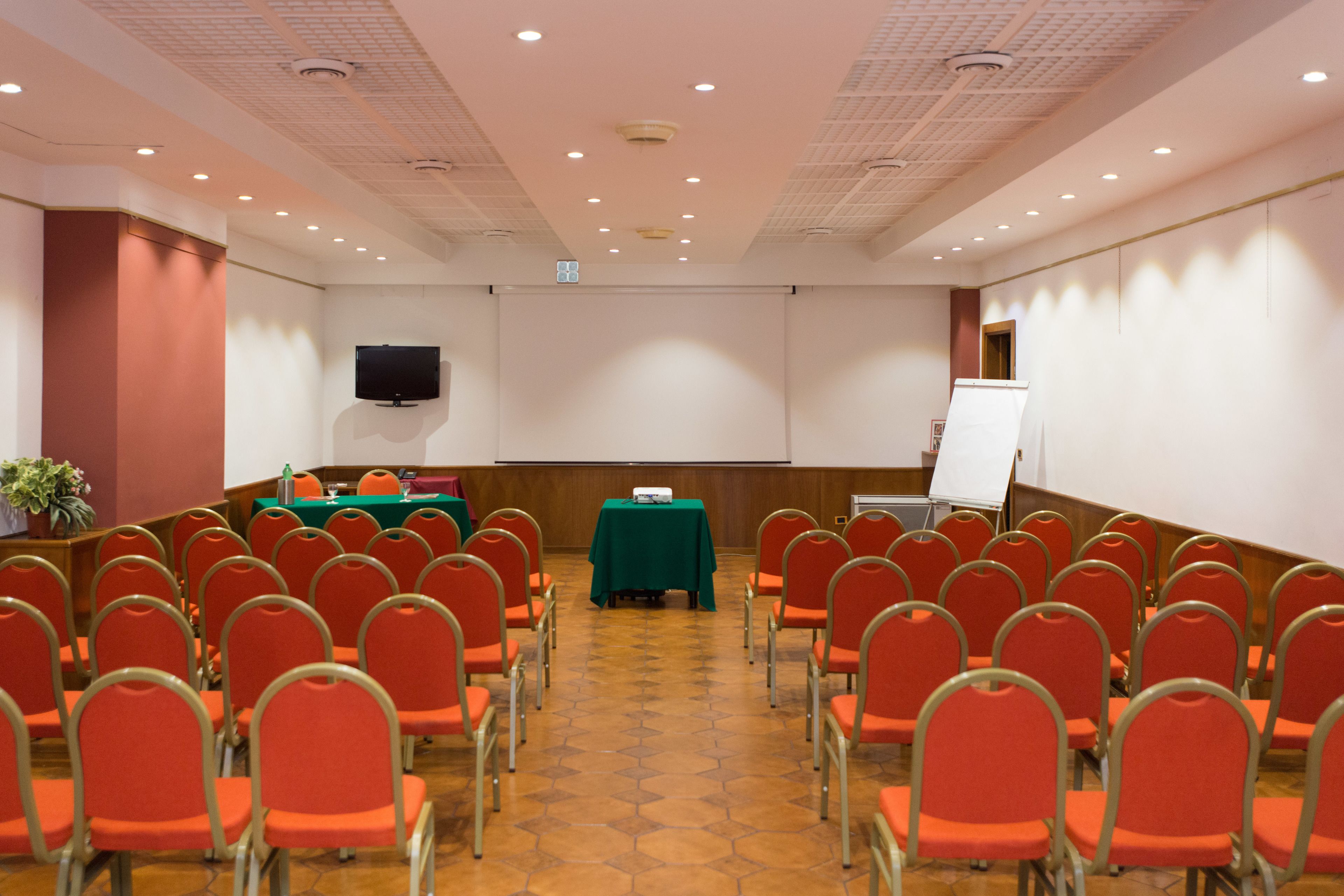 A conference room with rows of red chairs, a green table, a projector screen, and a television on the wall.