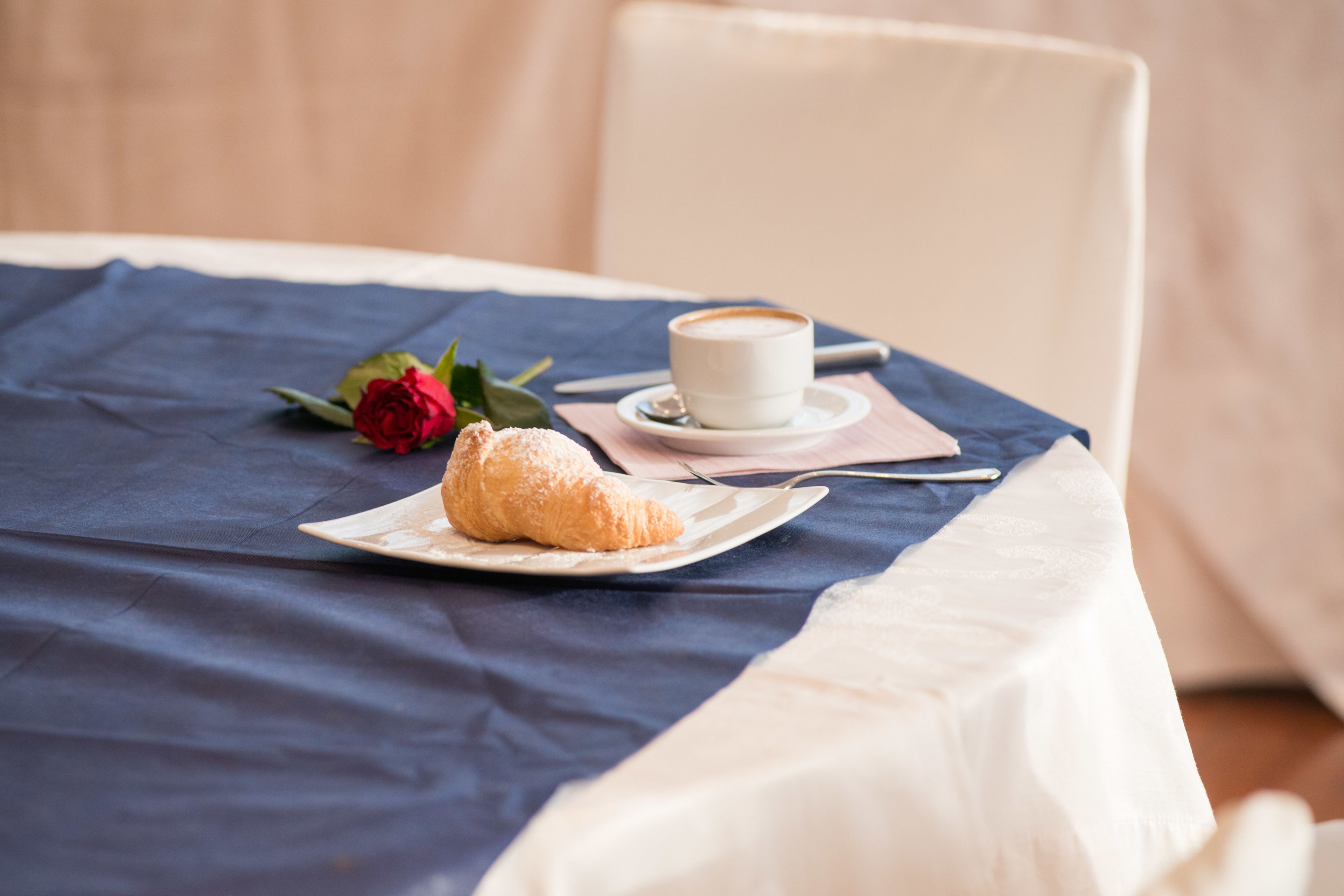 A table with a croissant, a cup of coffee, and a rose on a blue tablecloth.