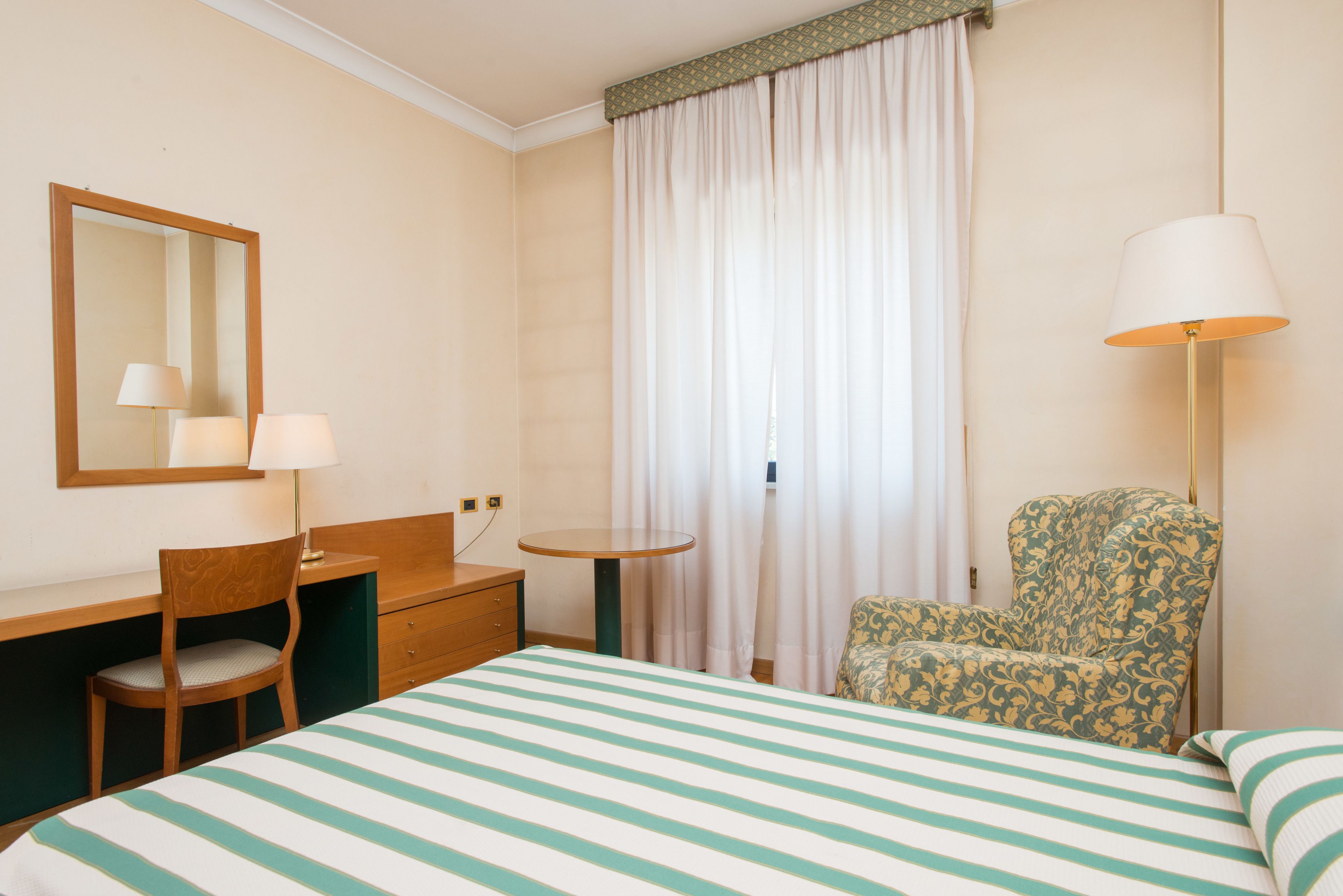 Una habitación de hotel con cama, escritorio, silla, lámpara y ventana con cortinas.