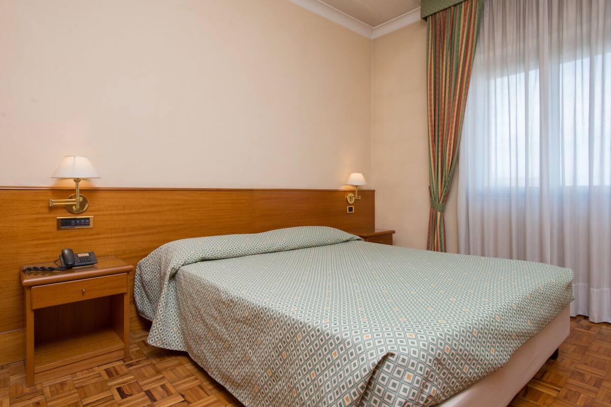 Une chambre d’hôtel avec une tête de lit en bois, deux lampes de chevet, un téléphone, une couvre-lit à motifs et une fenêtre avec des rideaux.
