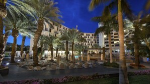 Un patio de hotel con palmeras, sillas de descanso y una zona de piscina al anochecer.