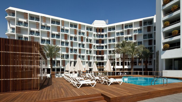 Un hotel moderno con varios balcones, una zona de piscina con tumbonas y una terraza de madera.
