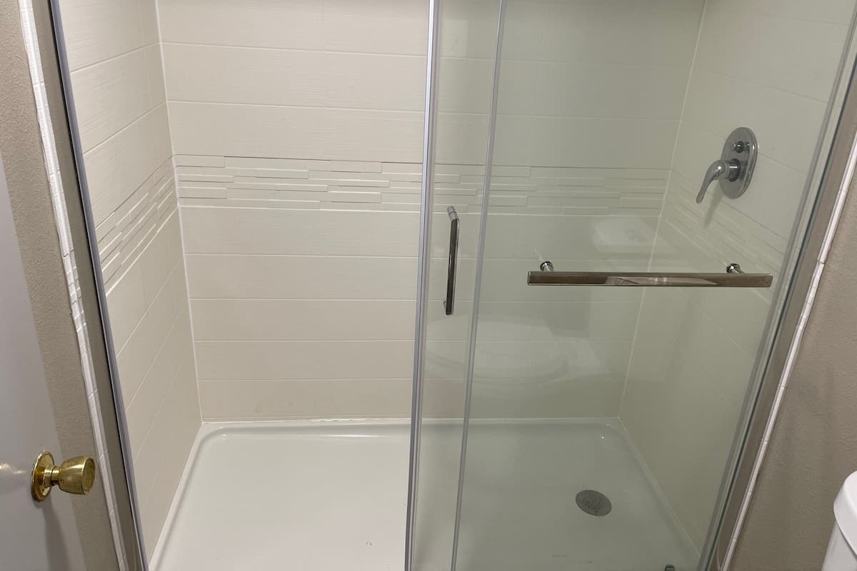 A glass-enclosed shower stall with a white tiled wall, a showerhead, and a grab bar.
