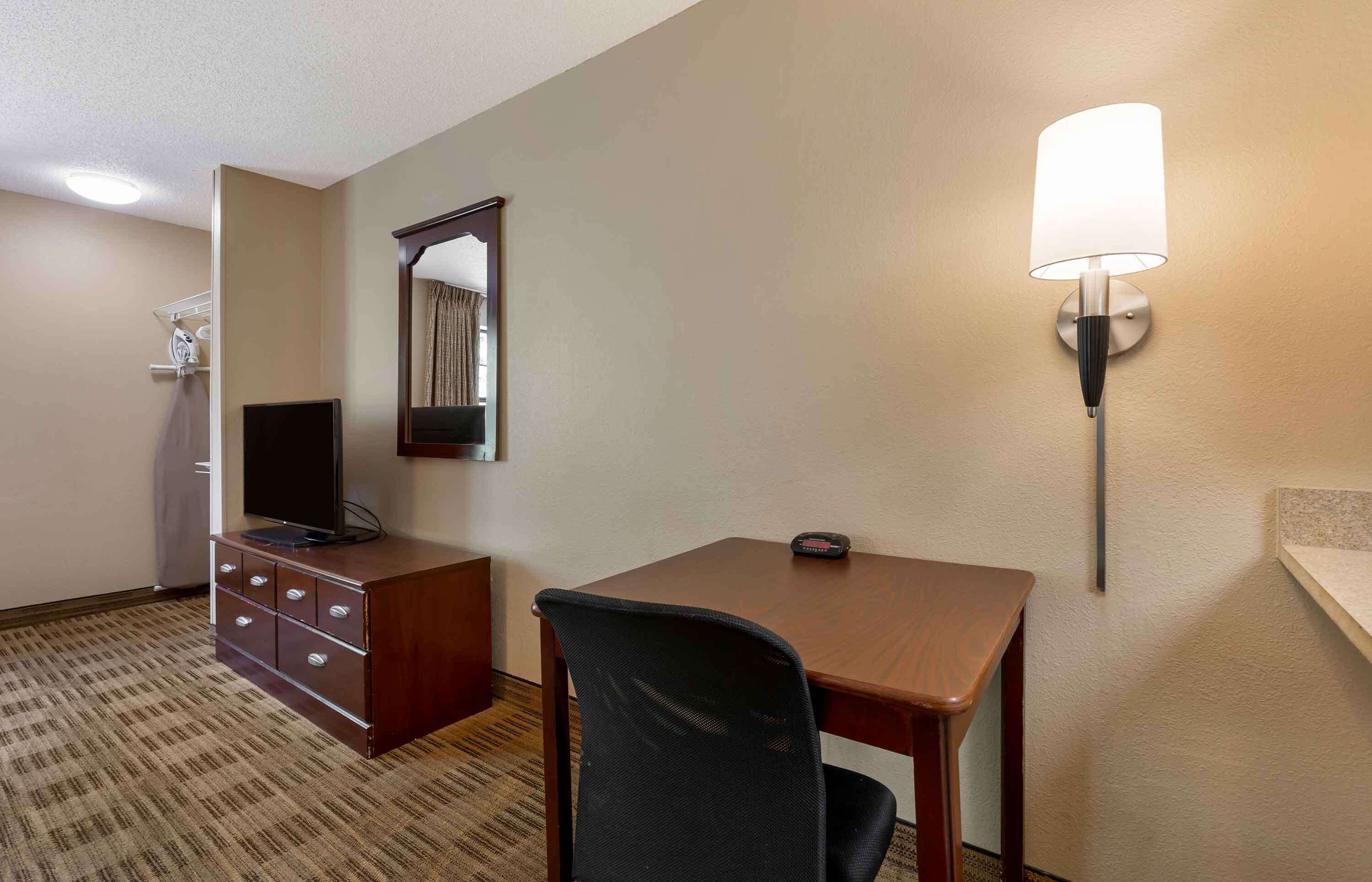 Habitación de hotel con un escritorio de madera, una silla de oficina negra, un televisor sobre una cómoda, una lámpara de pared y un espejo.