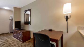 A hotel room with a wooden desk, a black office chair, a television on a dresser, a wall-mounted lamp, and a mirror.