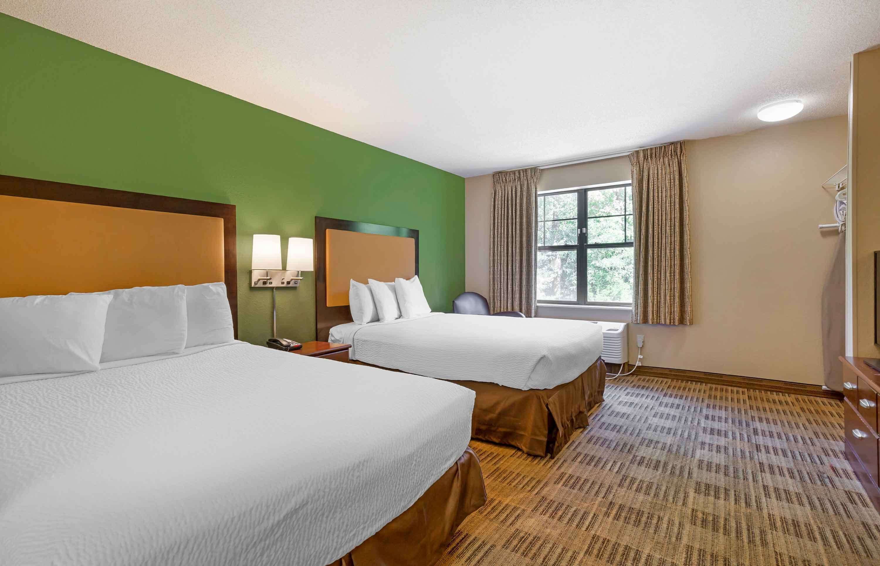 A hotel room with two beds, a large window with curtains, a desk with a lamp, and a television.