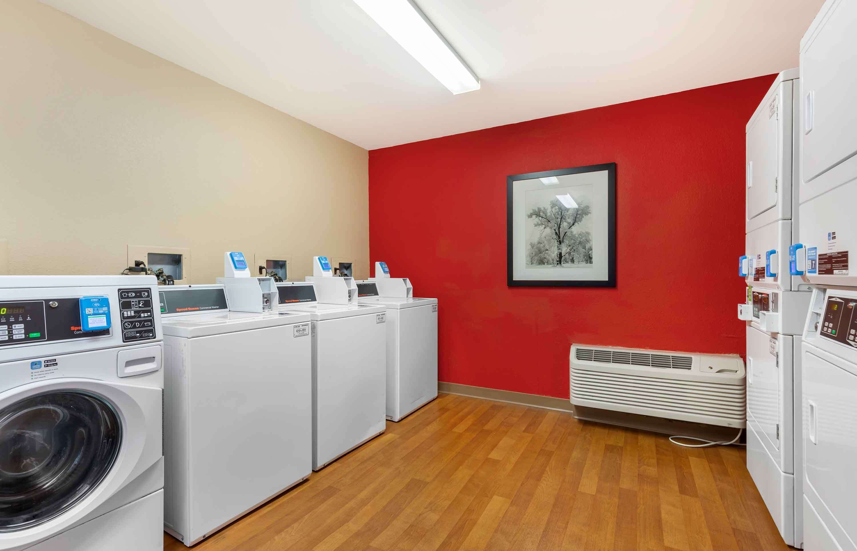 Une buanderie équipée de plusieurs machines à laver et de plusieurs sèche-linge, avec un mur rouge et un tableau encadré.