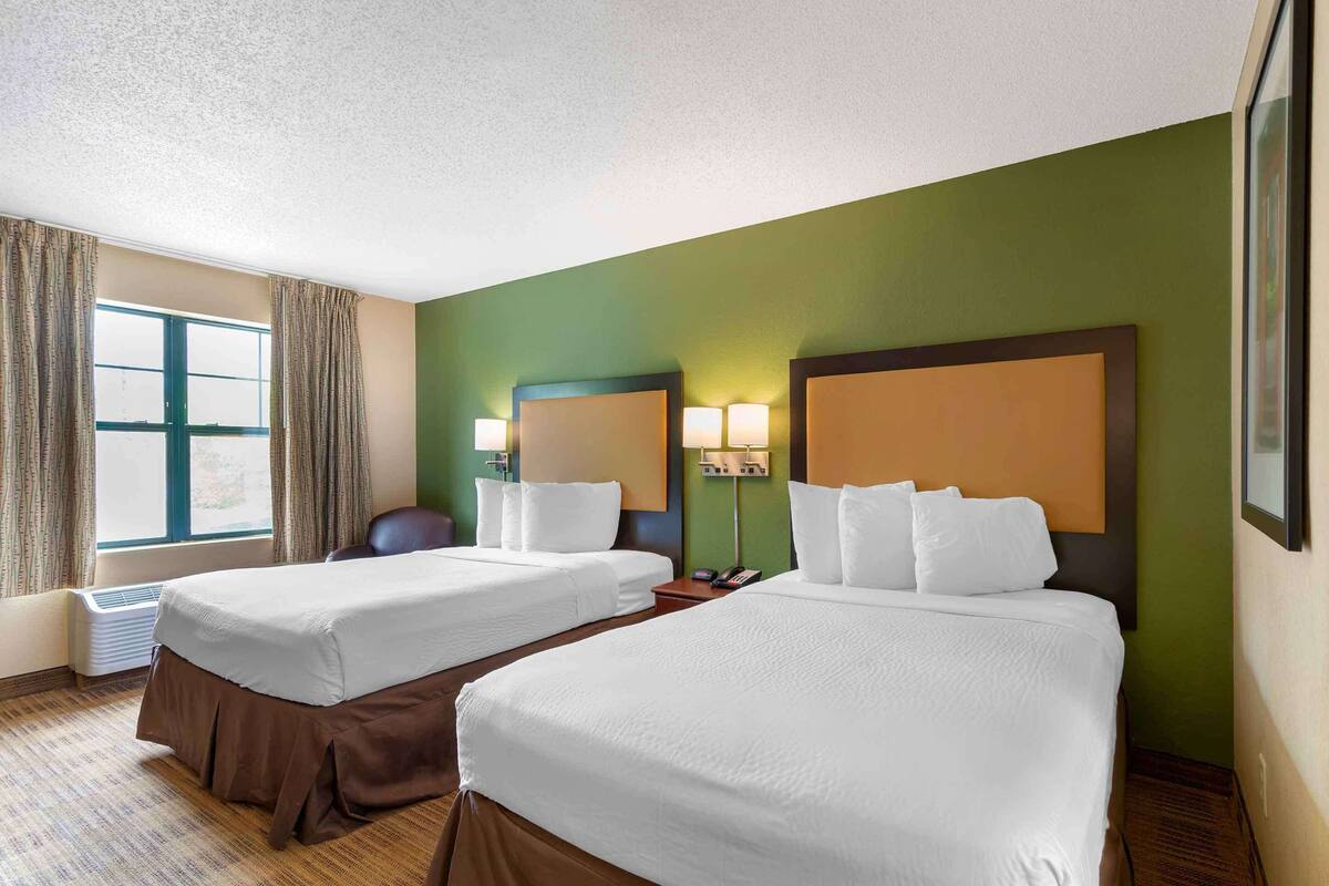 A hotel room with two beds, a large window with curtains, a desk with a lamp, and a wall-mounted phone.