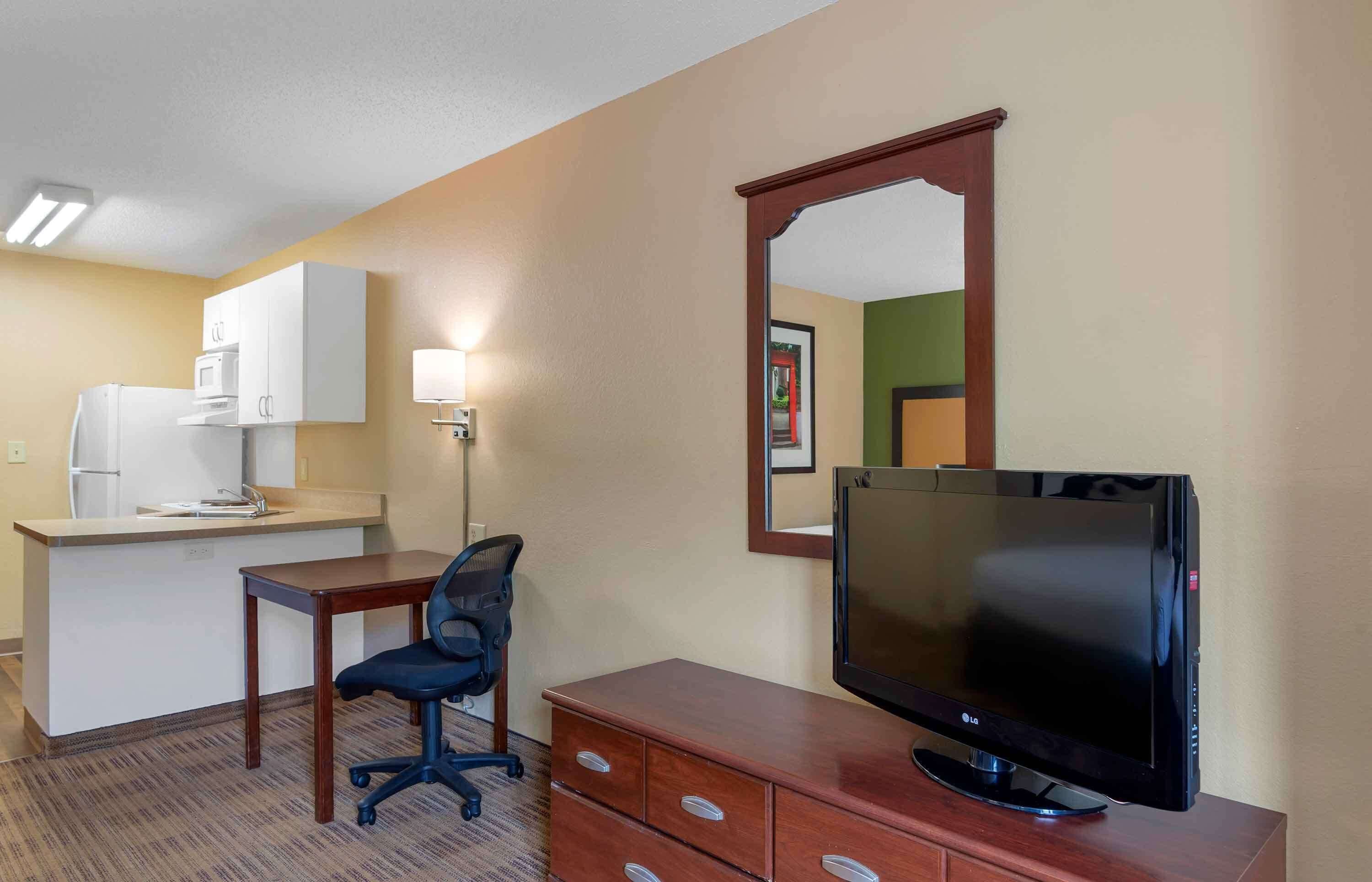 Ein Hotelzimmer mit Küchenzeile, einem Schreibtisch mit Stuhl, einem Flachbildfernseher und einem Spiegel.