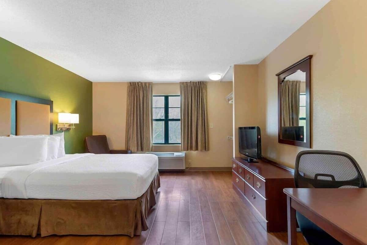 A hotel room with a bed, a desk, a chair, a television, a window with curtains, and a wall-mounted mirror.