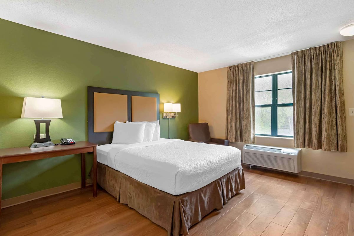 A hotel room with a bed, a desk with a lamp, a chair, a window with curtains, and an air conditioning unit.