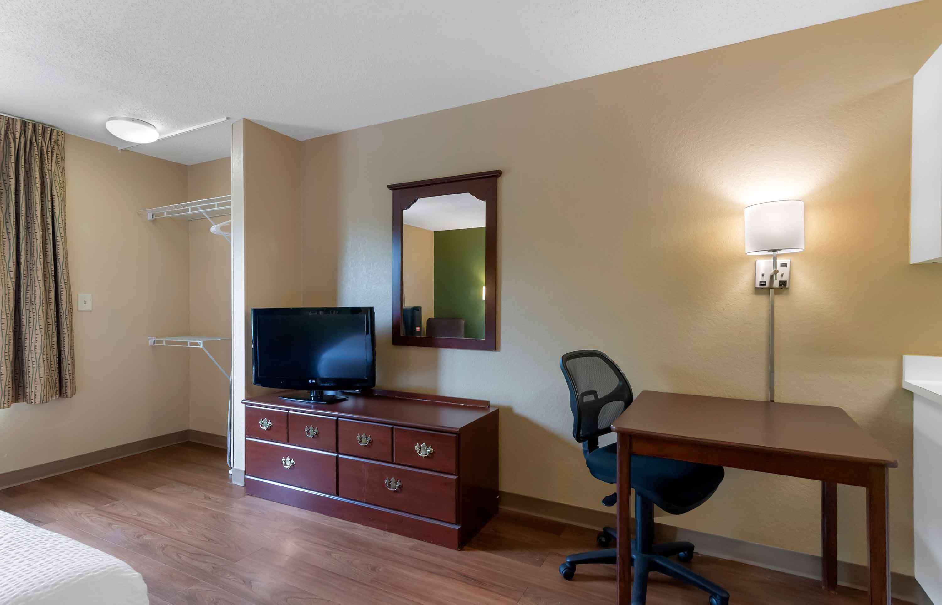 A hotel room with a wooden desk, a television on a dresser, a chair, a mirror, and a wall-mounted lamp.