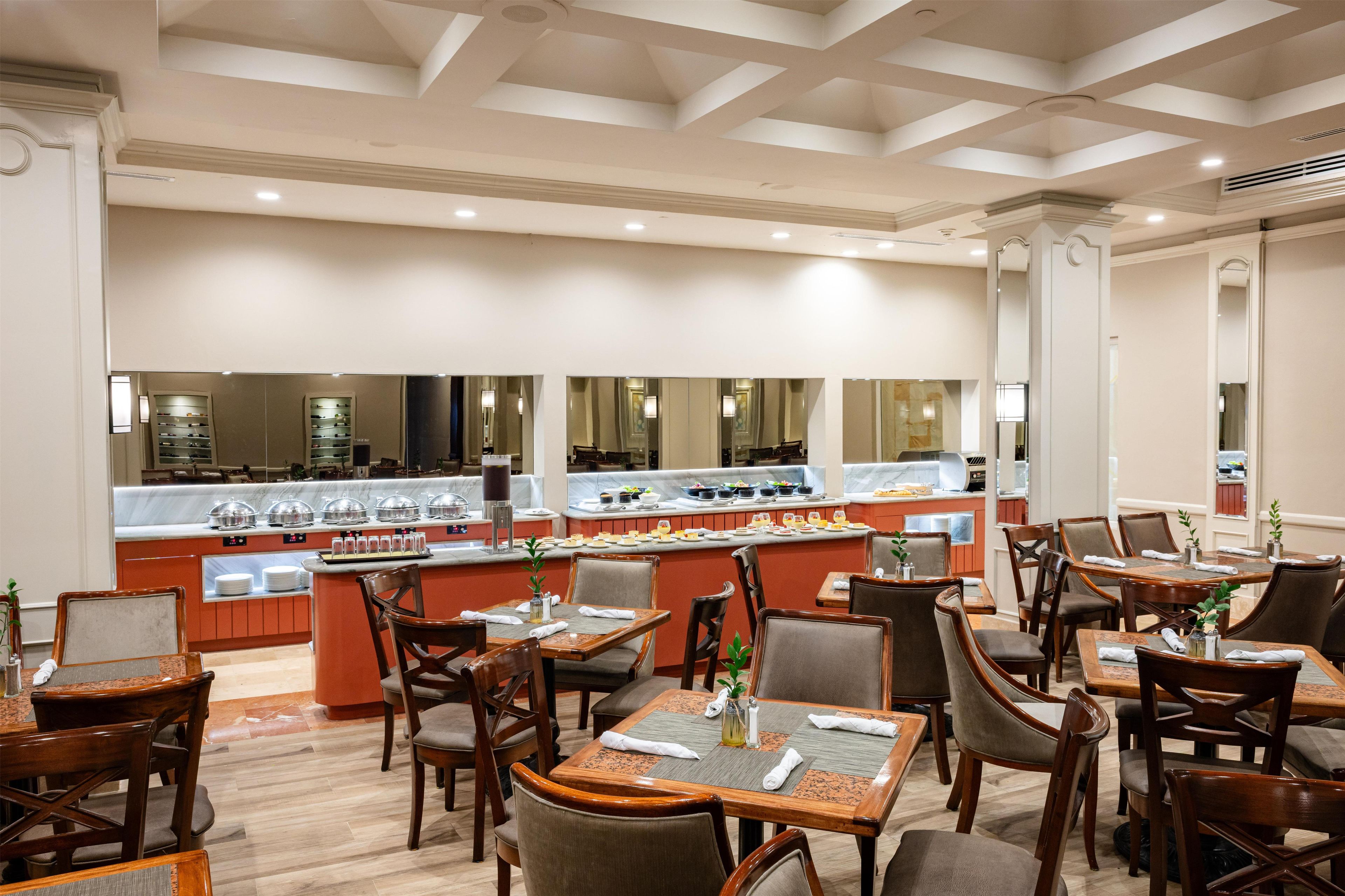 A hotel dining area with multiple tables and chairs, a buffet counter, and a large mirror reflecting the space.