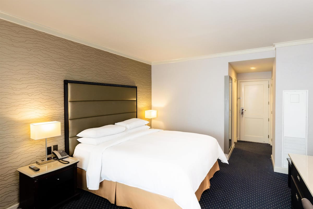 Ein Hotelzimmer mit Bett, Nachttisch mit Lampe, Telefon, Schrank und einer Tür zu einem weiteren Raum.