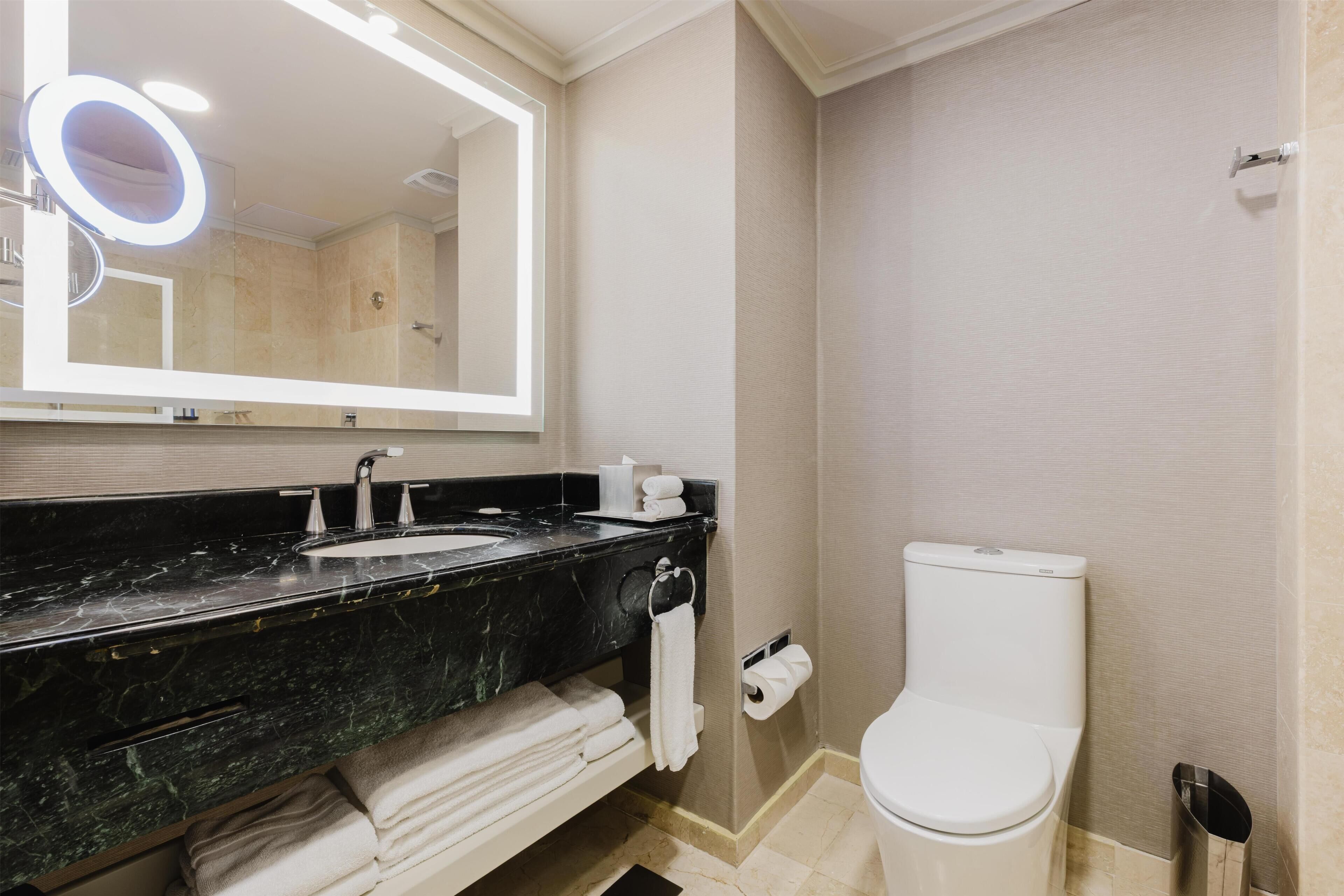 A bathroom with a marble sink, a toilet, a towel rack, and a mirror with a light.