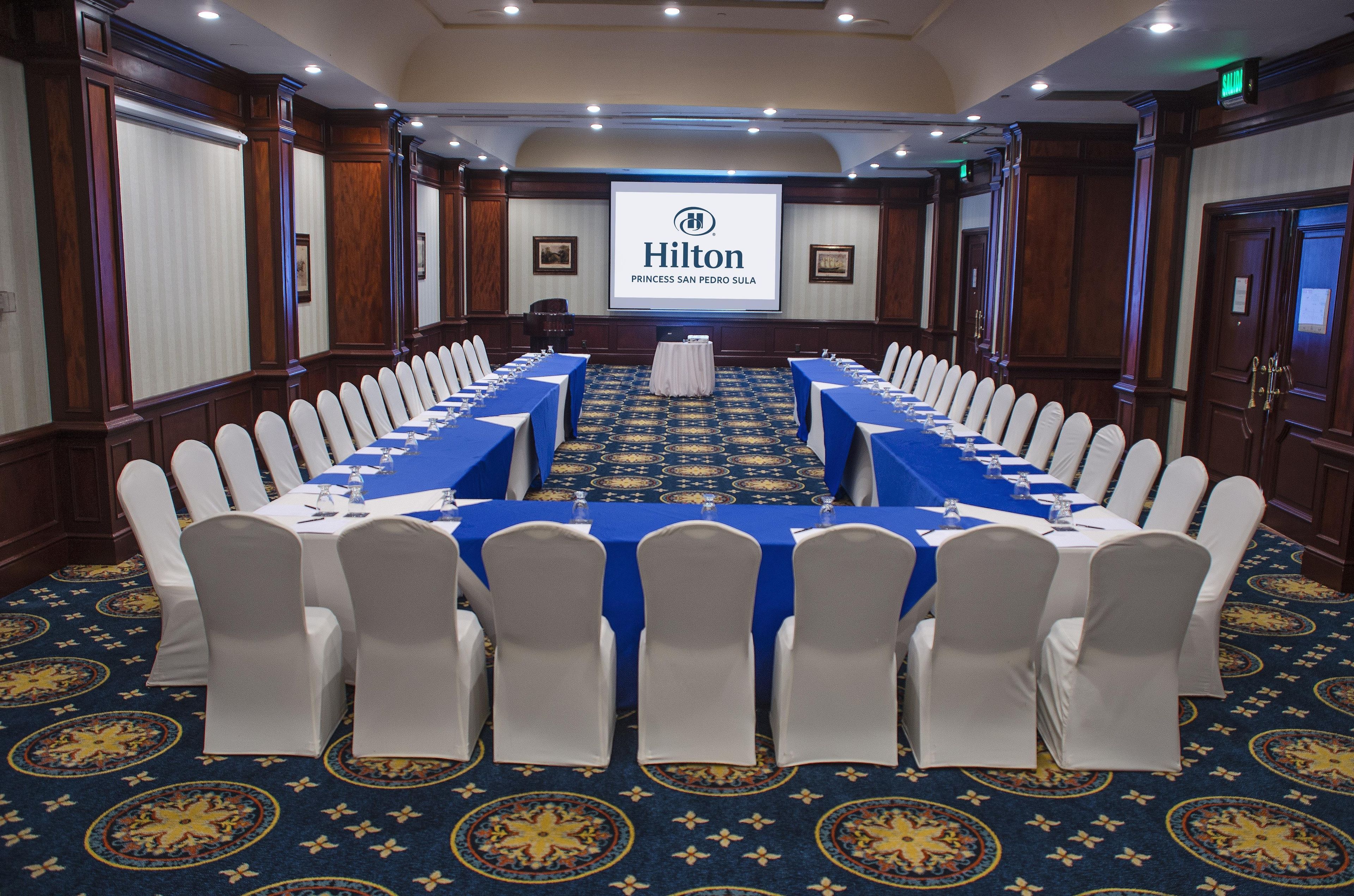 Une salle de conférence avec une longue table, des chaises blanches et un écran affichant le logo de Hilton.