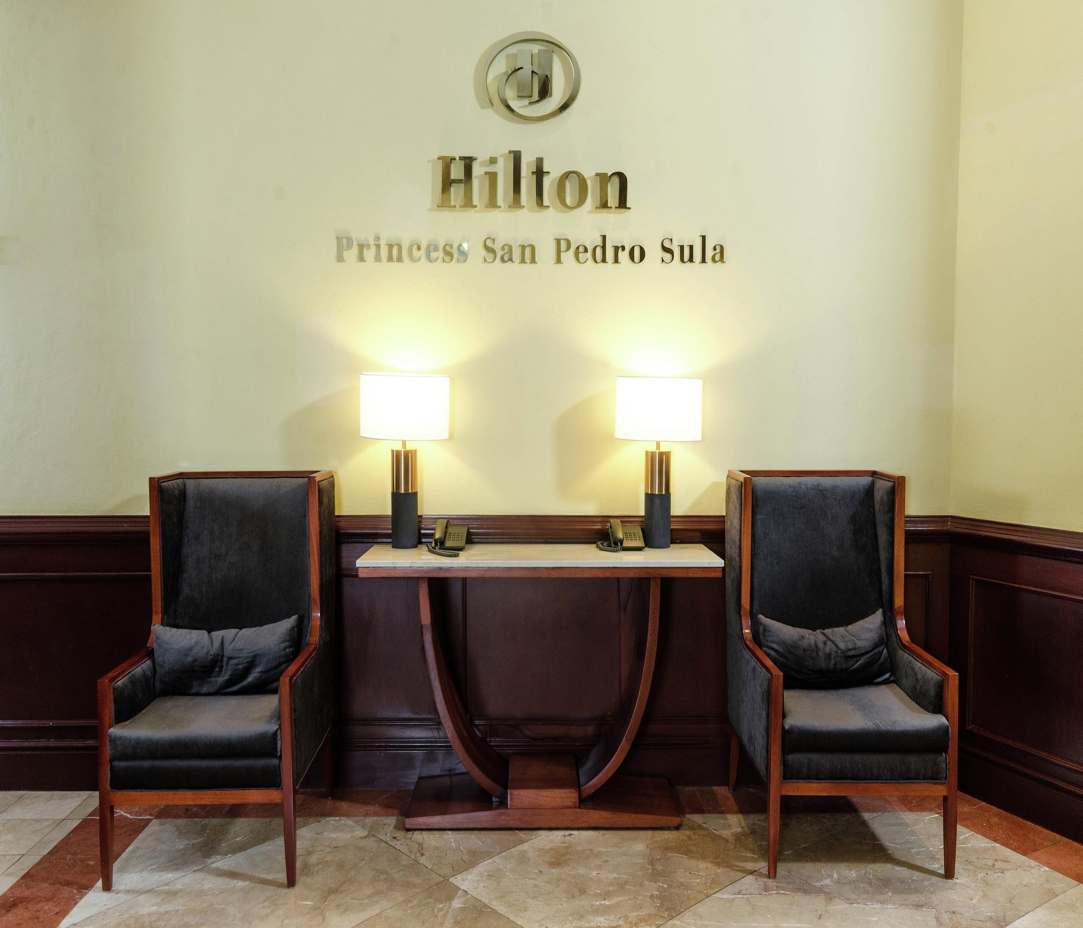 A reception area with two armchairs, a desk, and a wall-mounted sign reading 'Hilton Princess San Pedro Sula.'