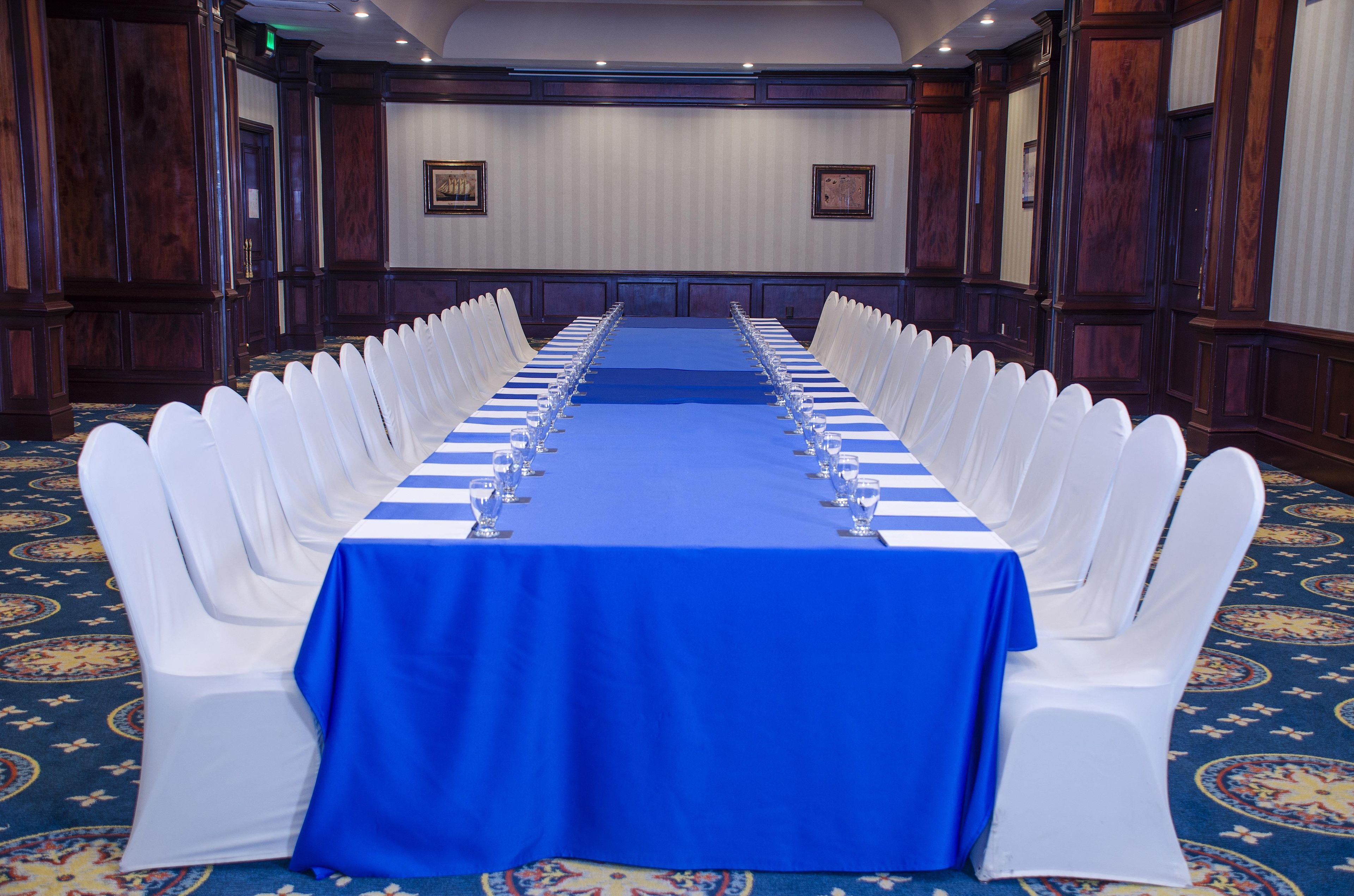 Un salón de conferencias con una larga mesa, sillas blancas y un mantel azul.