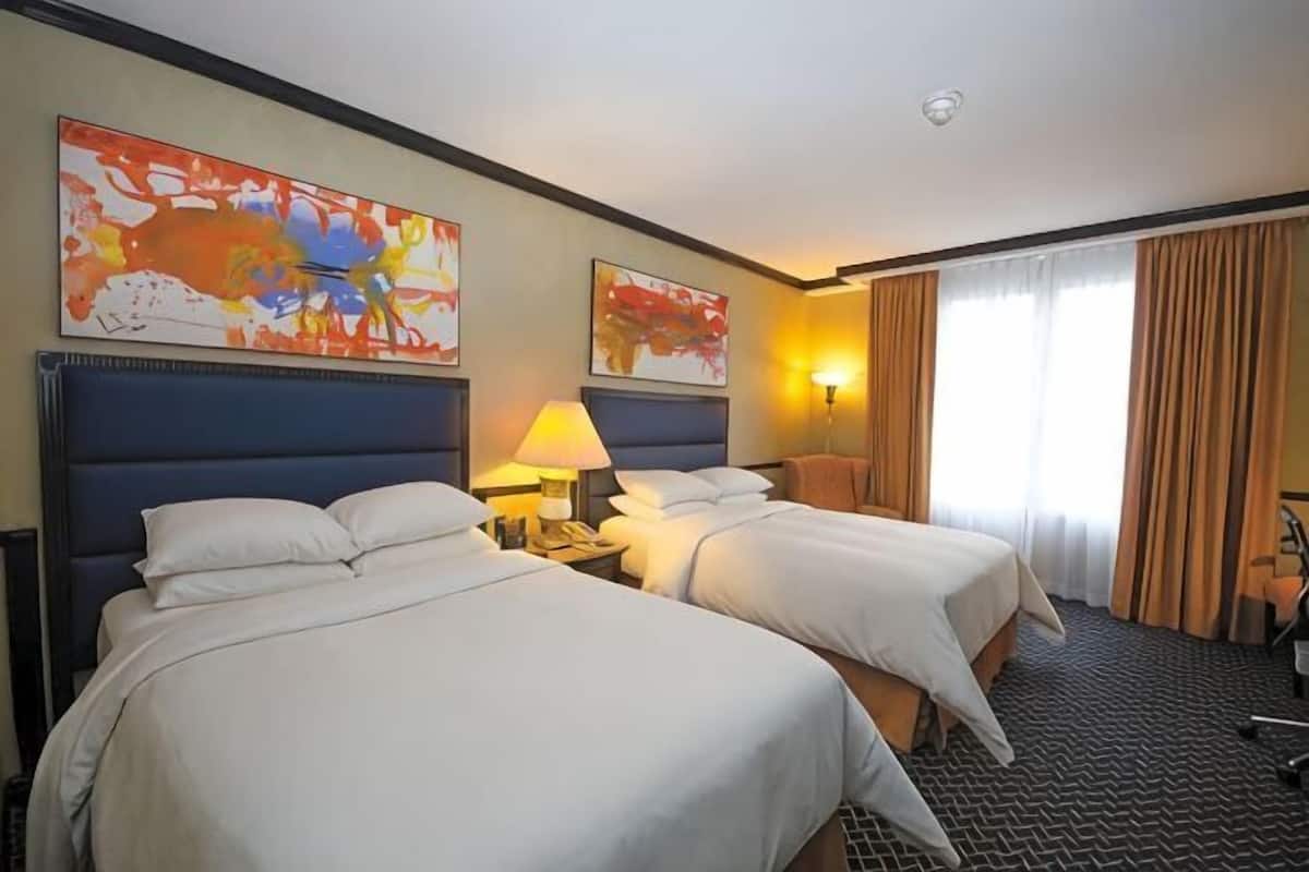 A hotel room with two beds, a desk, a lamp, and artwork on the walls.
