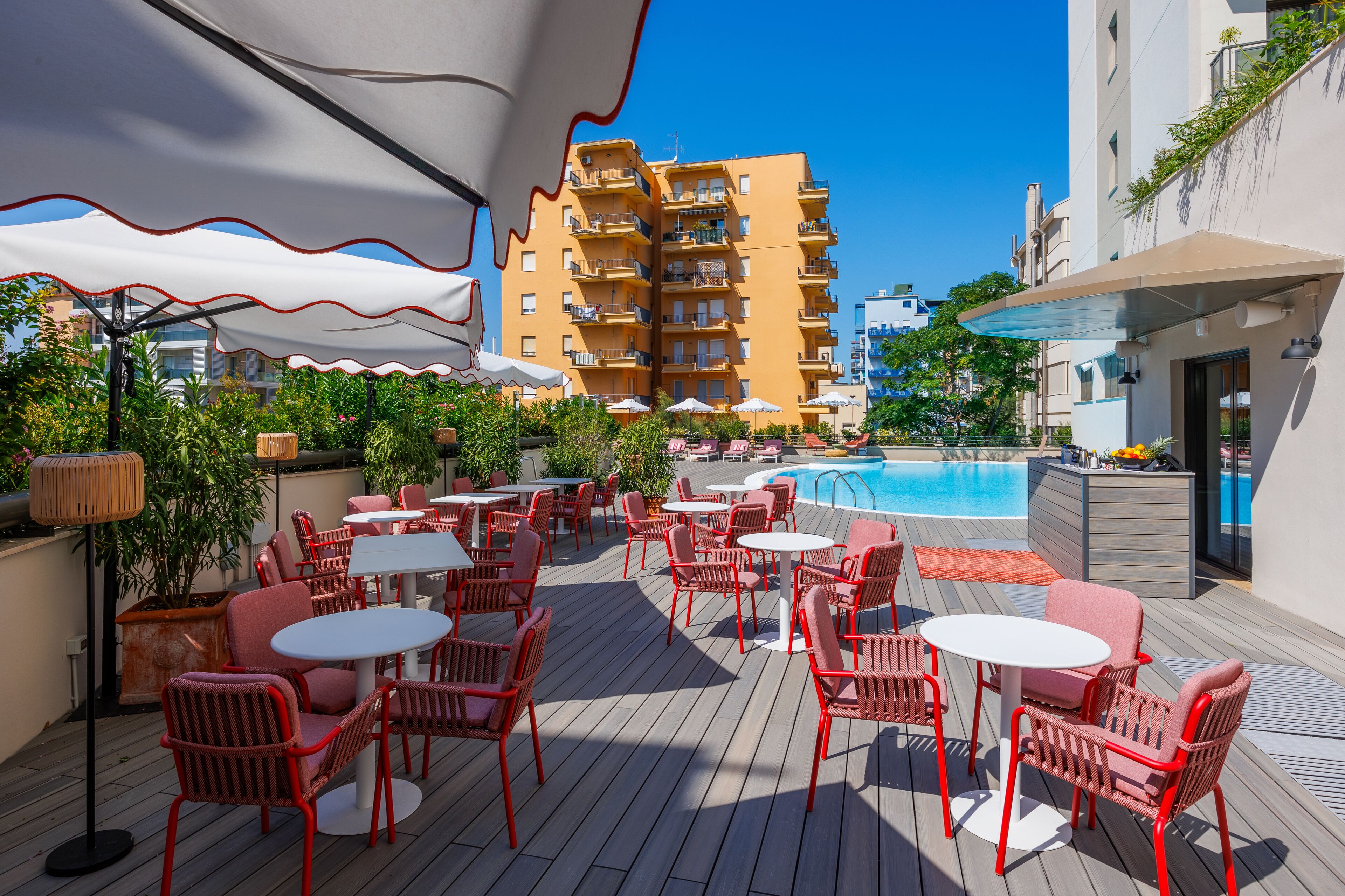 Una terraza en la azotea con mobiliario exterior, una piscina y un edificio de varios pisos al fondo.