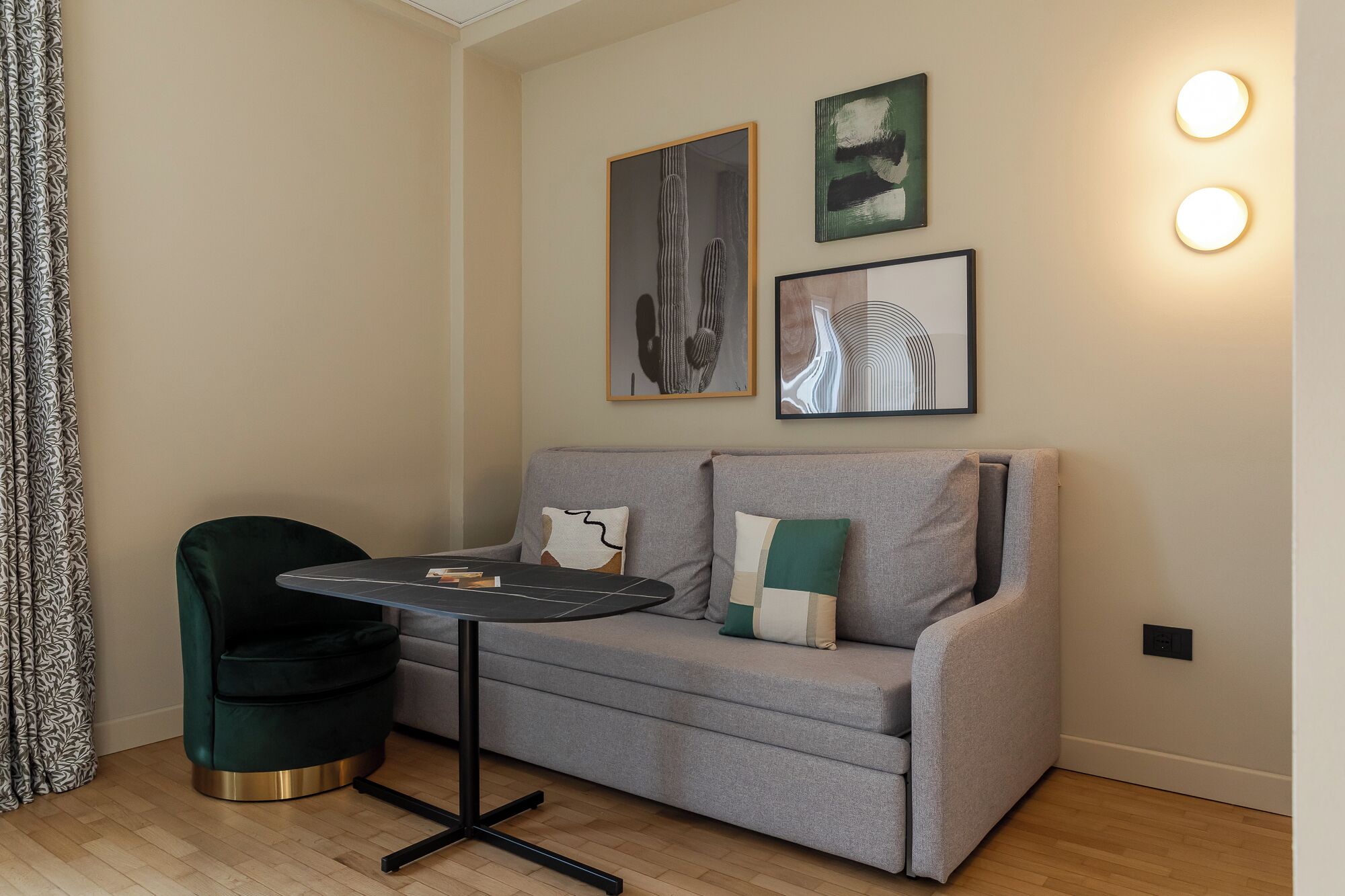A modern living room with a grey sofa, a green armchair, and a small black table. There are two framed artworks on the wall and a wall-mounted light fixture.