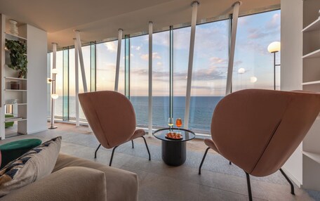 Un salón moderno con ventanas amplias, dos sillones, una mesa de centro y una estantería.