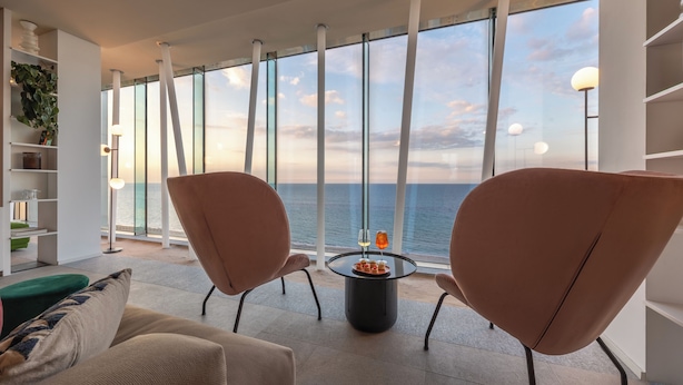 Un salón moderno con ventanas amplias, dos sillones, una mesa de centro y una estantería.