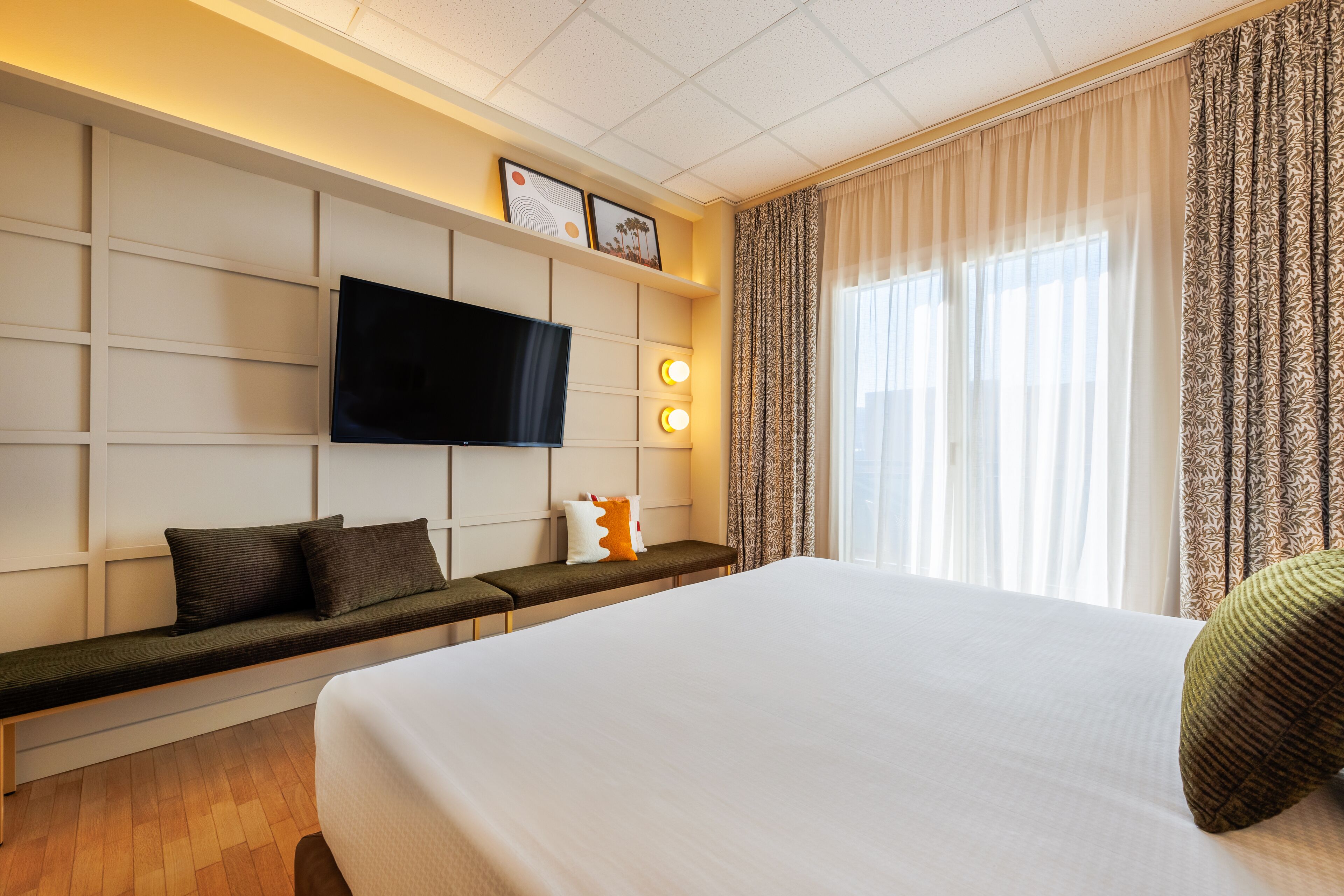 A hotel room with a large bed, a flat-screen TV mounted on the wall, a bench with pillows, and a window with curtains.