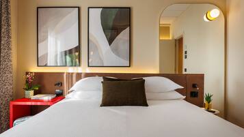 Una habitación de hotel con una cama grande, una mesita de noche roja, un espejo fijado a la pared y una obra de arte abstracta.