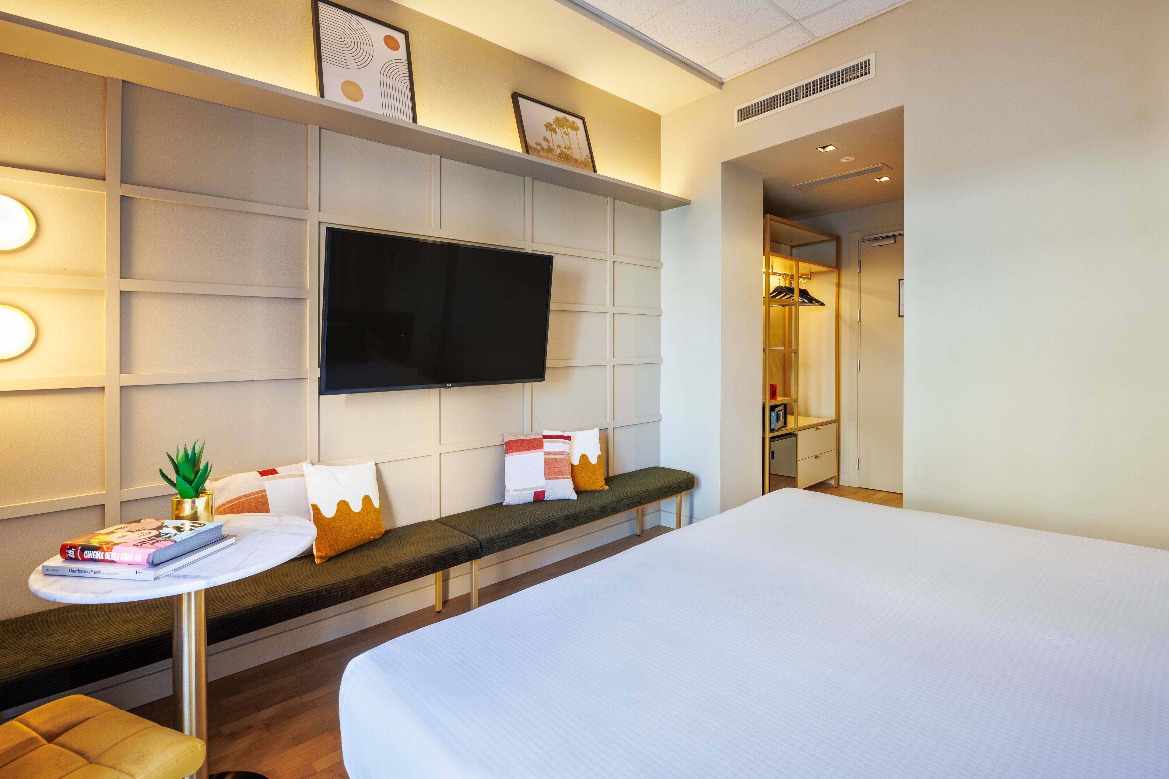 A hotel room with a large bed, a flat-screen TV mounted on the wall, a small round table with a plant, and a bench with cushions.